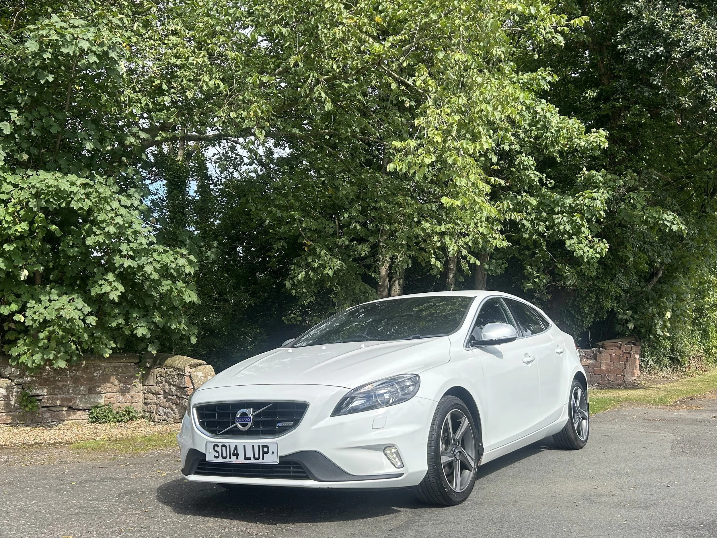 White Volvo car parked on a street with green trees and brick wall in the background.