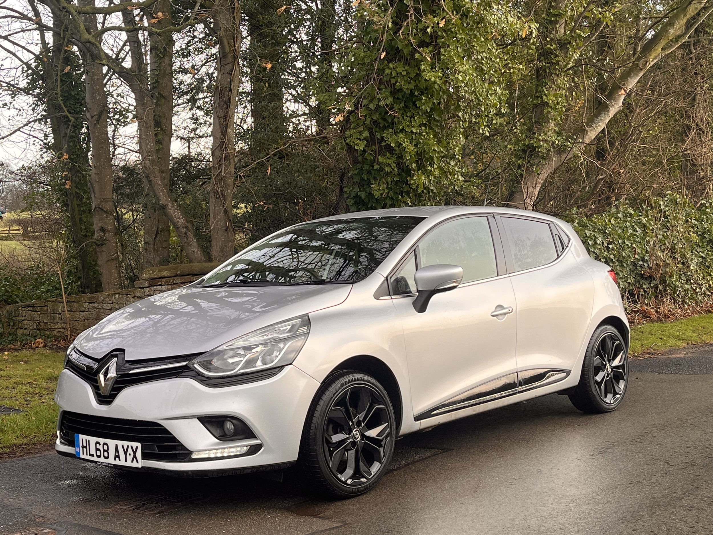 A silver Renault car parked on the side of a road next to a grassy area and trees