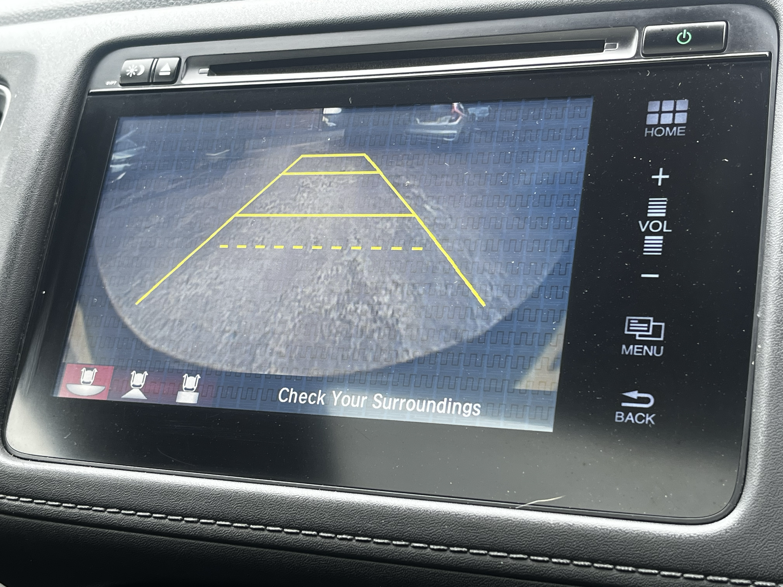 Car rearview camera display showing parking guidelines over a parking lot surface with the message "Check Your Surroundings" at the bottom of the screen.