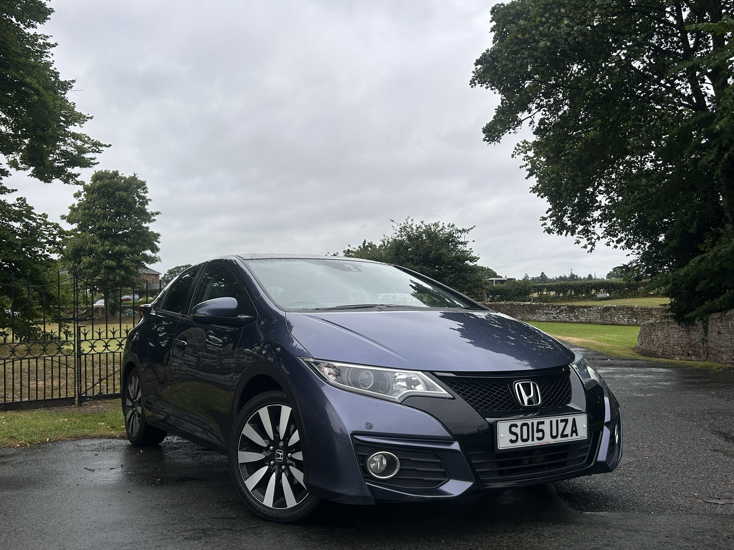 A black Honda Civic parked on a wet road, showing the front and side of the car. Behind the car is a black iron fence, green grass, trees, and a cloudy sky.