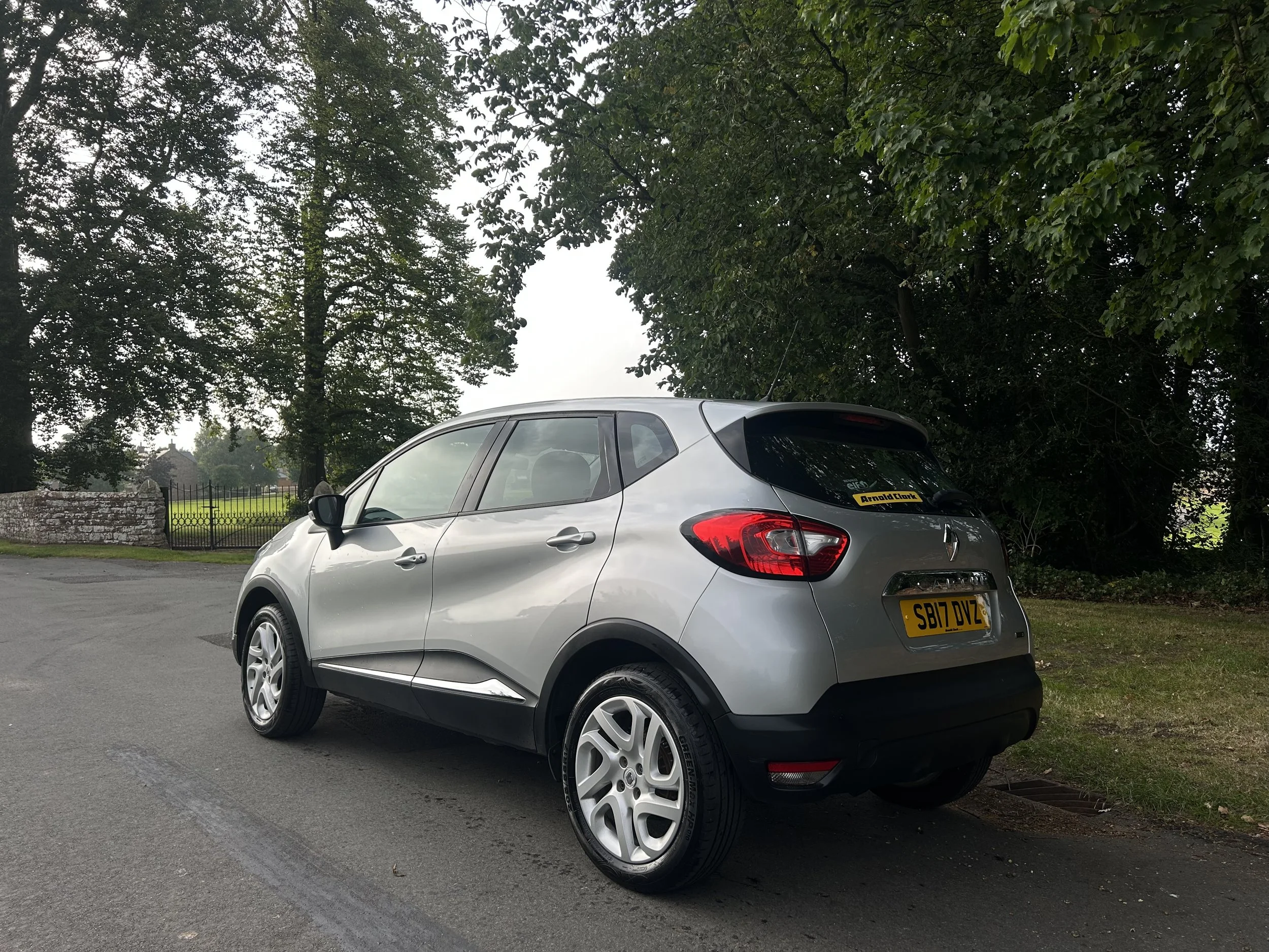 Silver Renault Captur parked on the street near trees and a grass area with a stone wall and black gate in the background.