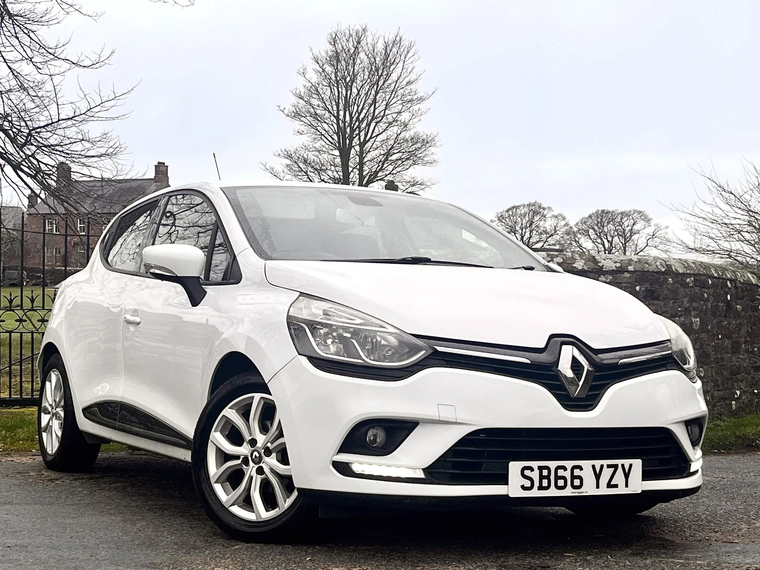 White Renault hatchback car parked outdoors on a cloudy day with leafless trees and some buildings in the background