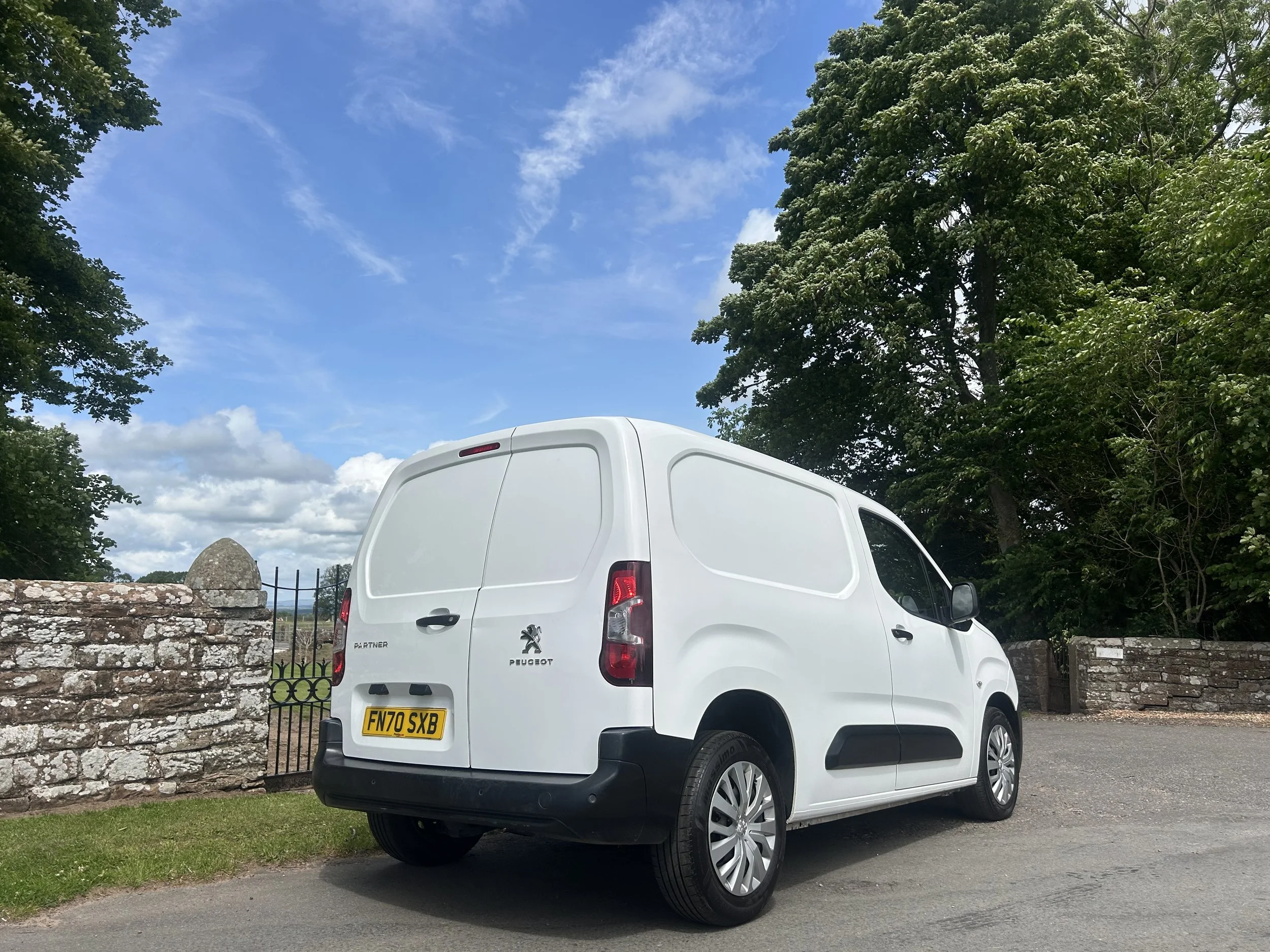 White Peugeot Partner cargo van parked on a paved surface with trees and a stone wall in the background under a blue sky with scattered clouds.