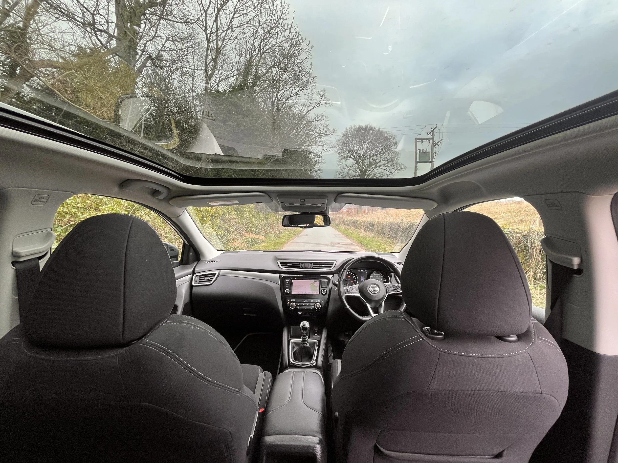 View of the interior of a car from the back seat, showing the dashboard, steering wheel, and two front seats. The car has a panoramic sunroof showing a cloudy sky and trees outside.