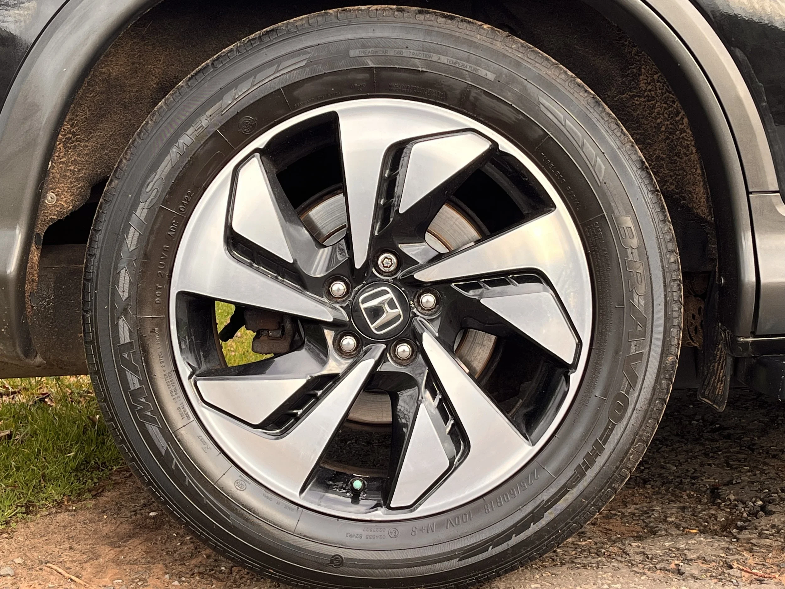 Close-up of a modern Honda car wheel with a black and silver alloy rim and a Bridgestone tire, showing part of the brake system and some rust around the wheel arch.