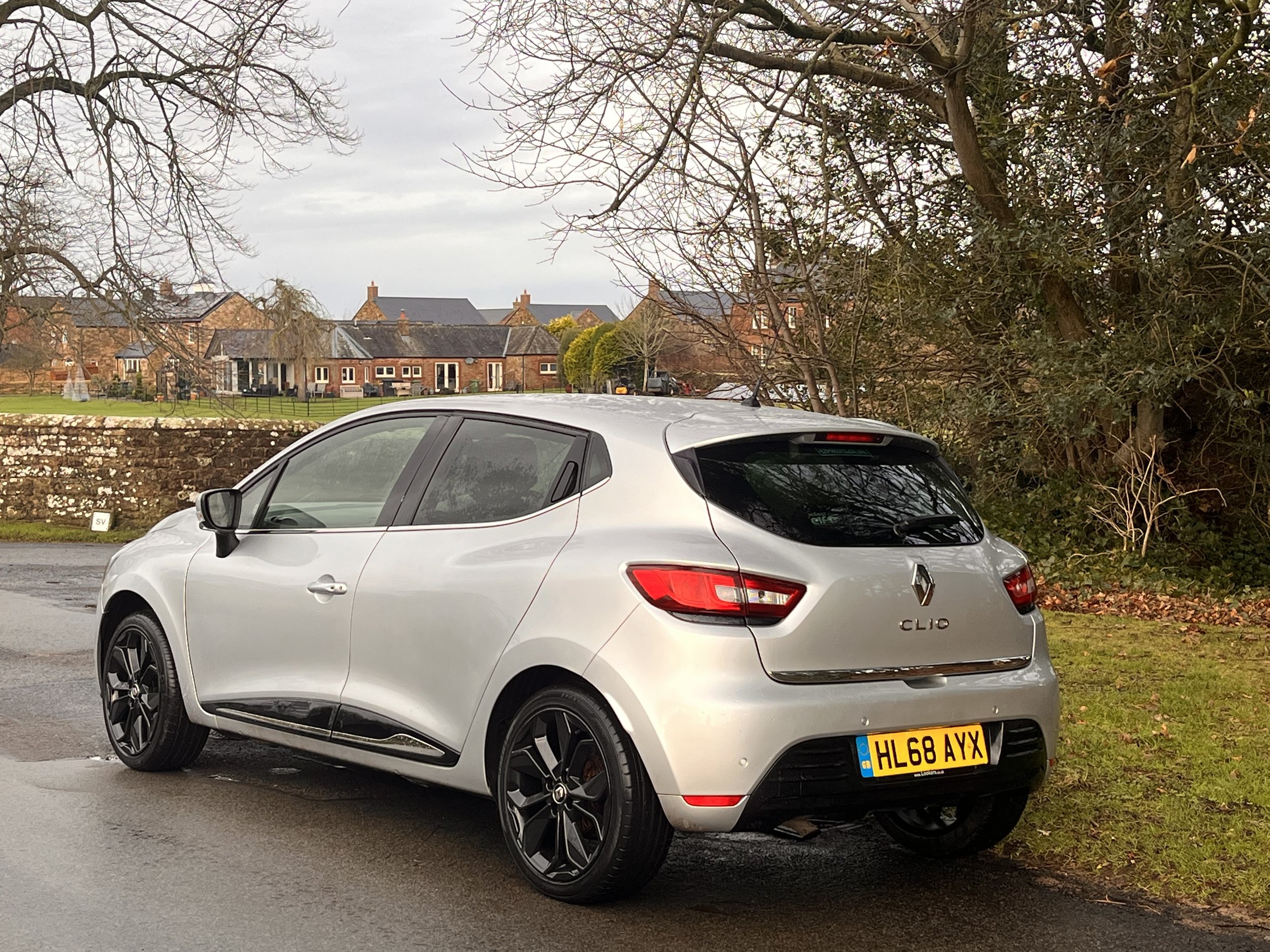 Silver Renault Clio parked on the side of a road near a grassy area with trees in the background.