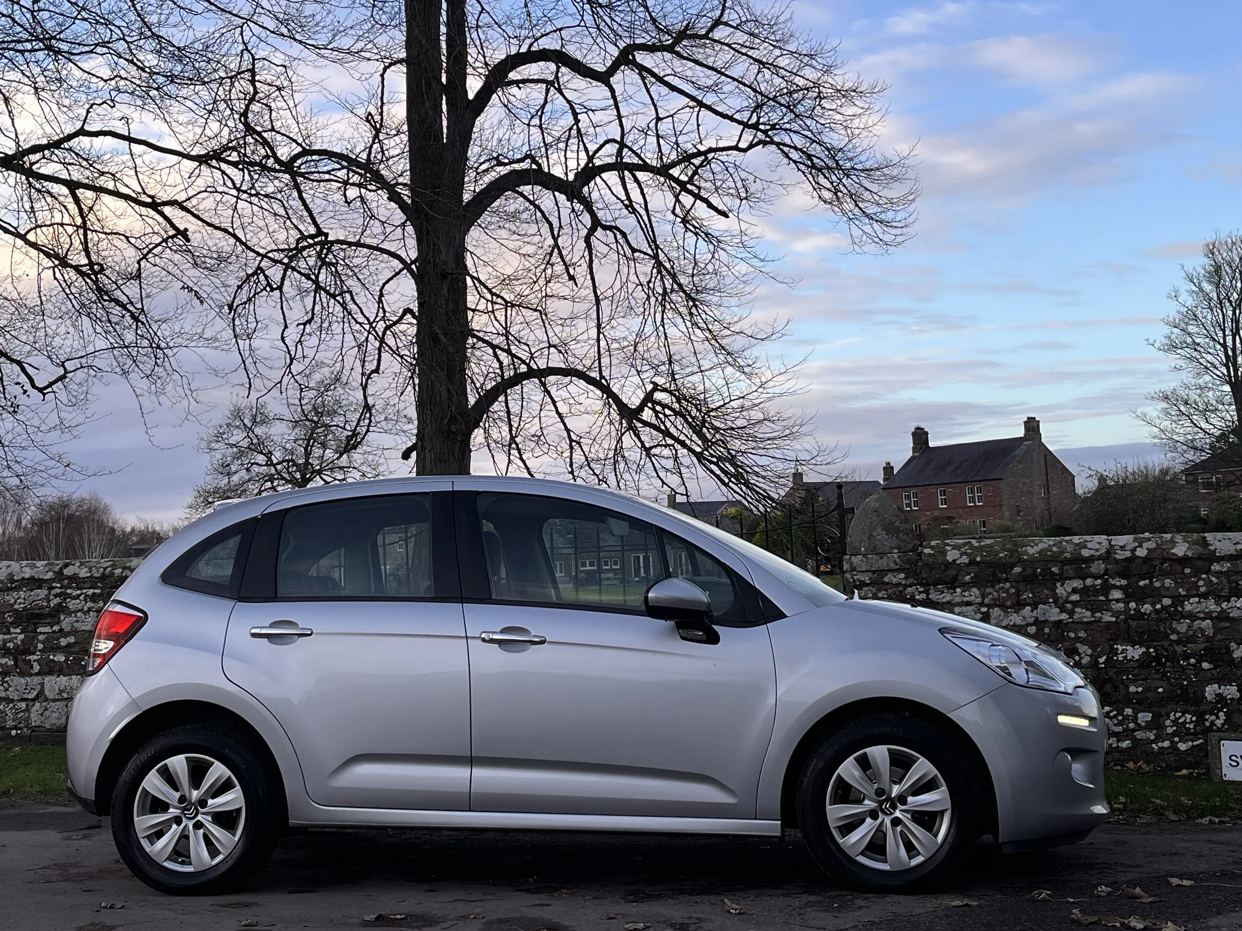 A silver compact hatchback car parked on the street with a stone wall, leafless tree, and houses in the background during dusk with a cloudy sky.