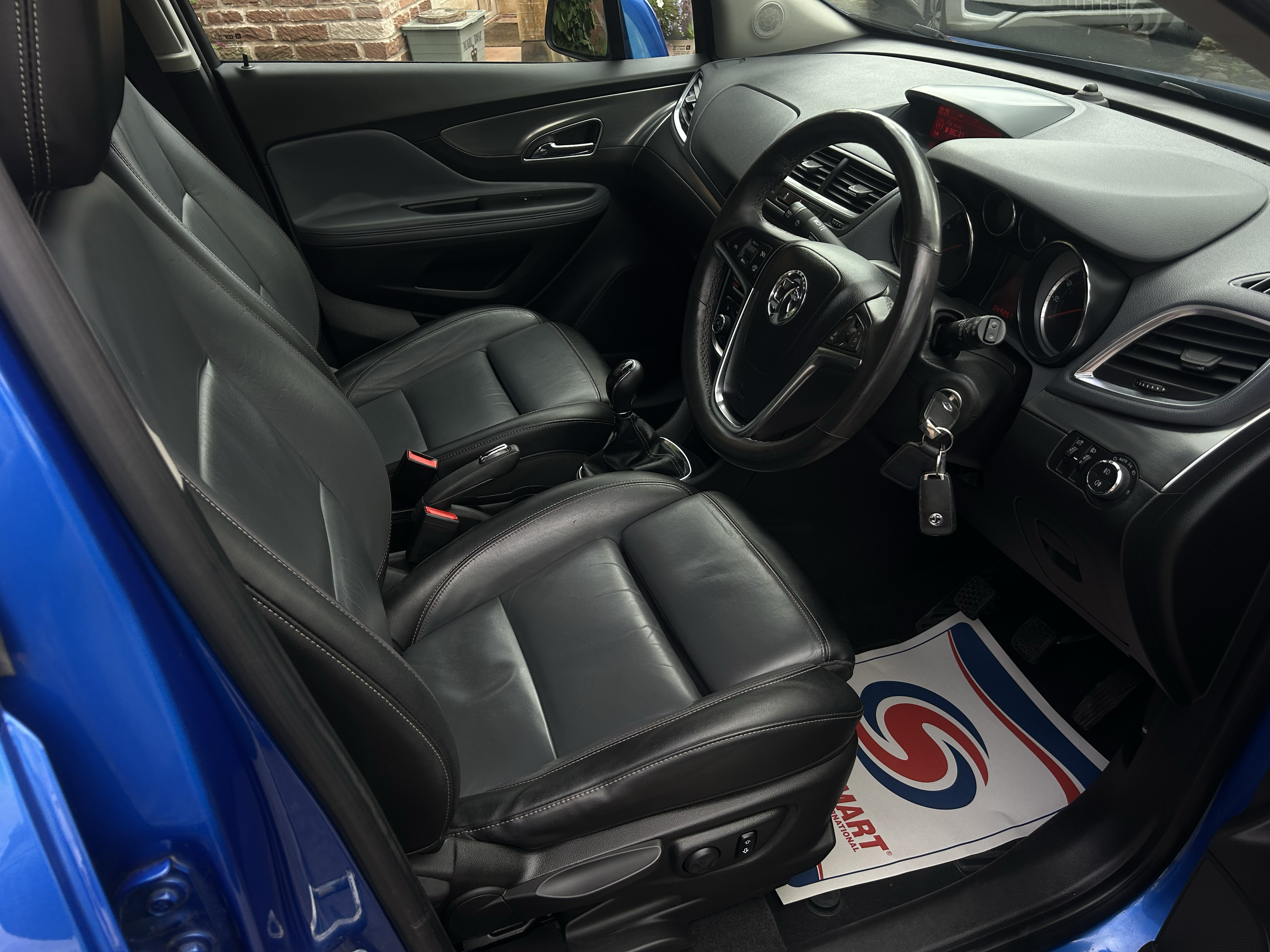 The interior of a car showing black leather seats, a steering wheel, gear shift, and dashboard with various controls, with brick wall and outdoor items visible through the window.