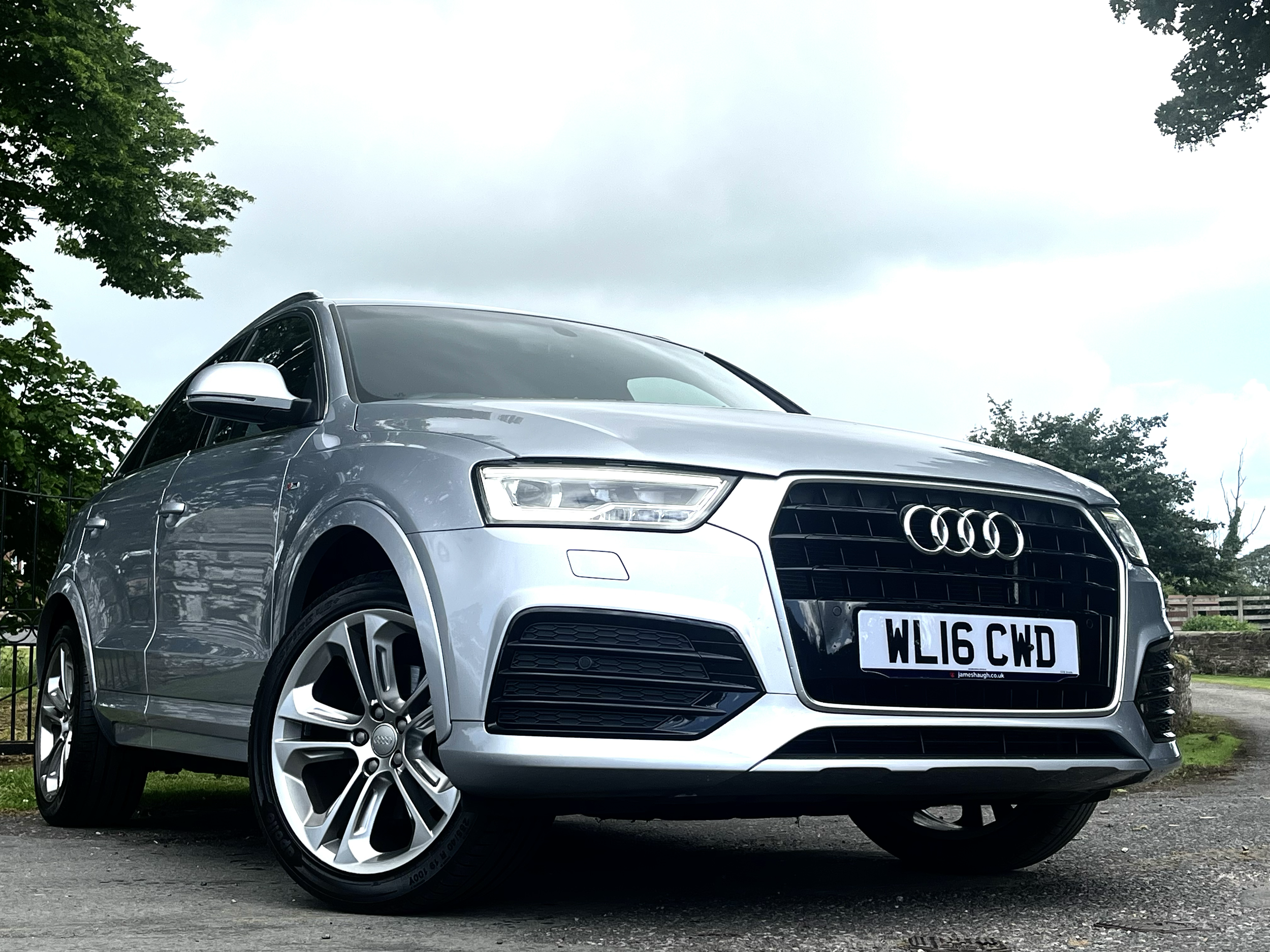 A silver Audi SUV parked outdoors on a paved surface with trees and a cloudy sky in the background.