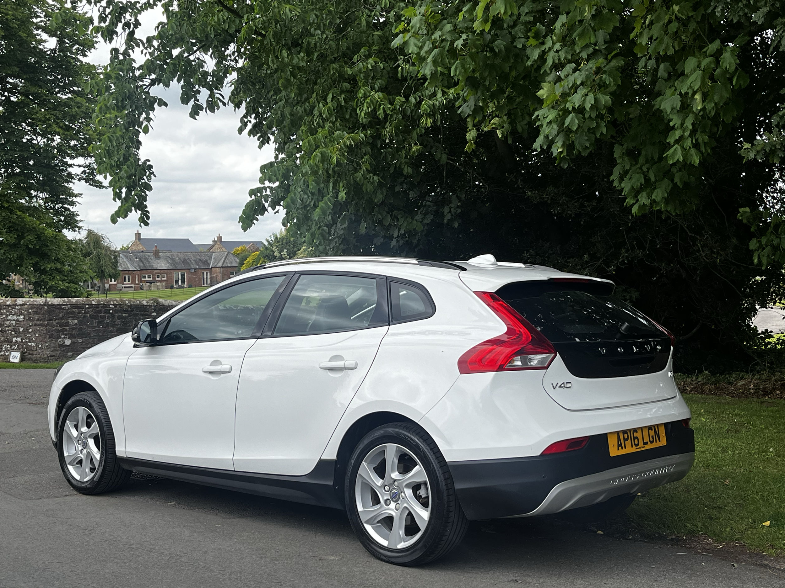 White hatchback car parked on a street with a stone wall behind it and green trees and a cloudy sky overhead.