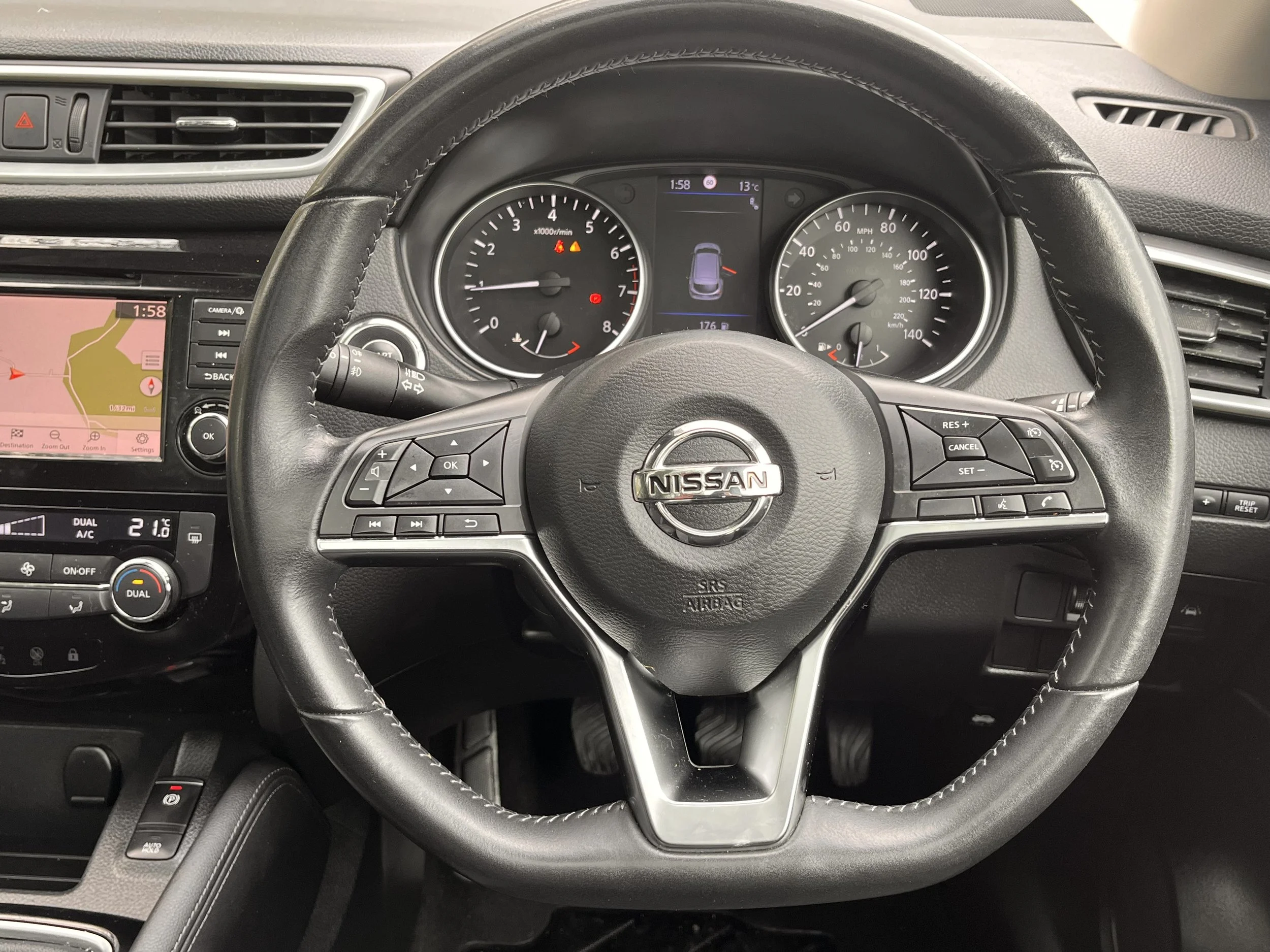 Inside view of a Nissan car dashboard and steering wheel. The speedometer, tachometer, and a digital display showing a small car icon and fuel range are visible. A GPS navigation screen and climate control panel are also seen.