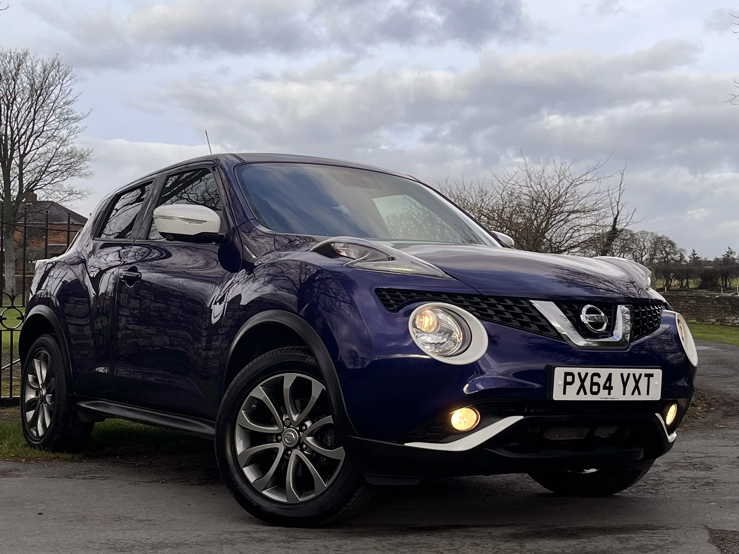 A purple Nissan Juke parked on a paved surface with a background of trees, a fence, and cloudy sky.