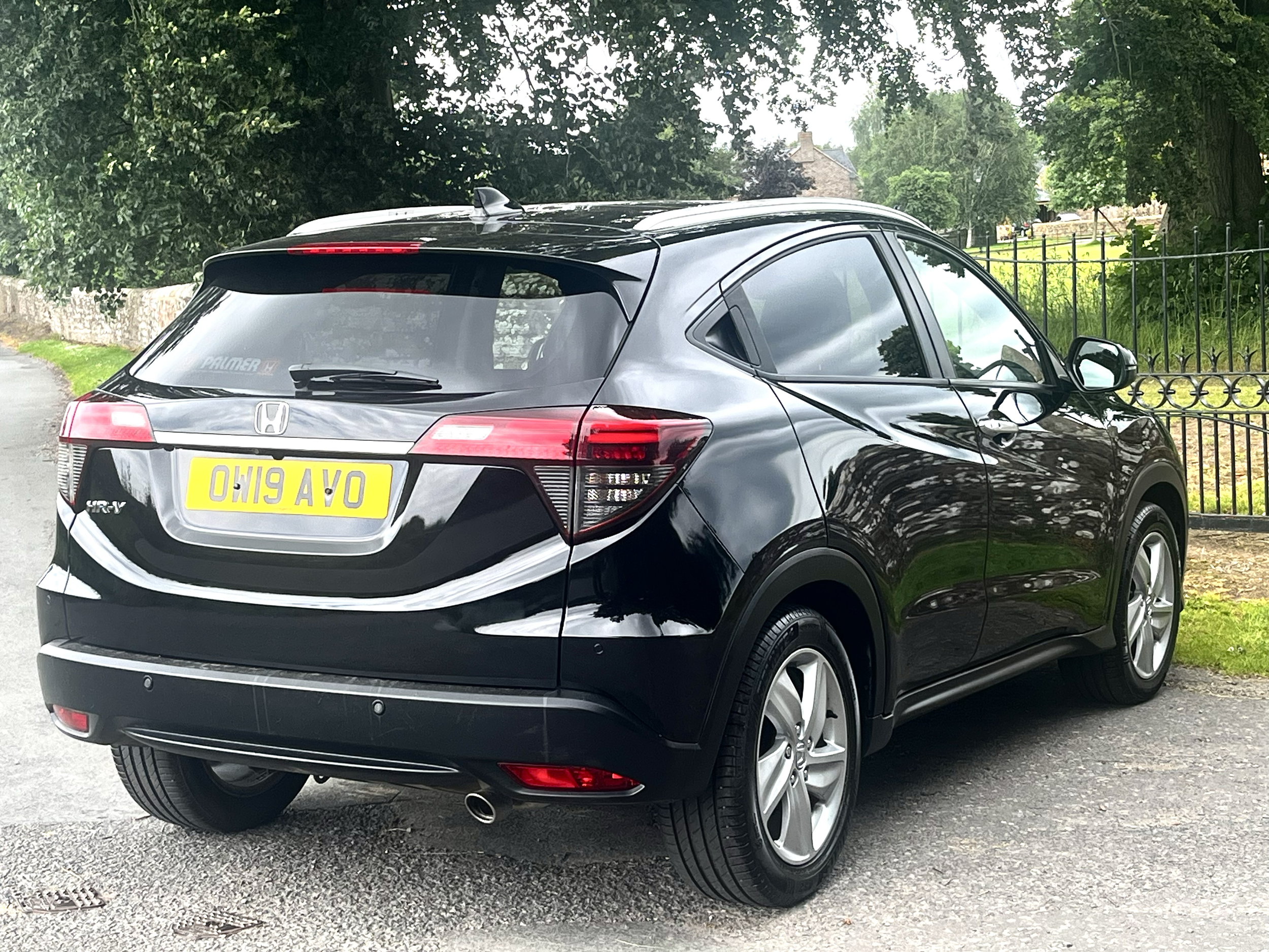 A black Honda HR-V parked on a gravel surface next to a black metal fence, with lush green trees and a stone building in the background.