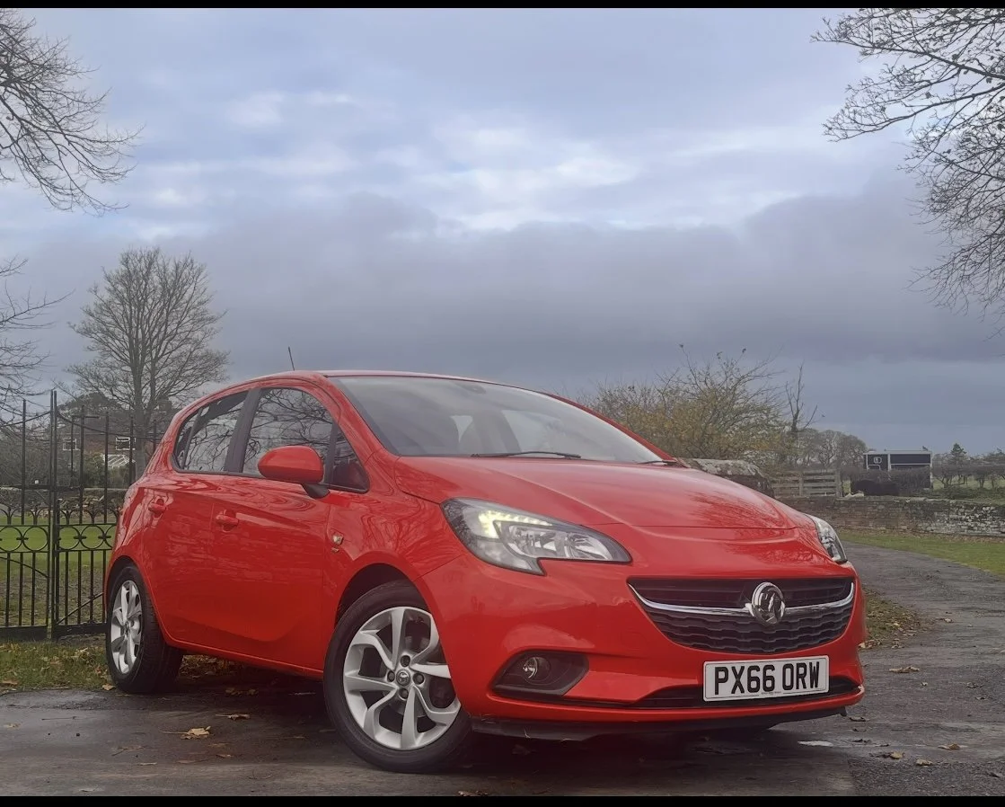 A red hatchback car parked outdoors on a paved surface with a rural background including trees, a fence, and cloudy skies.