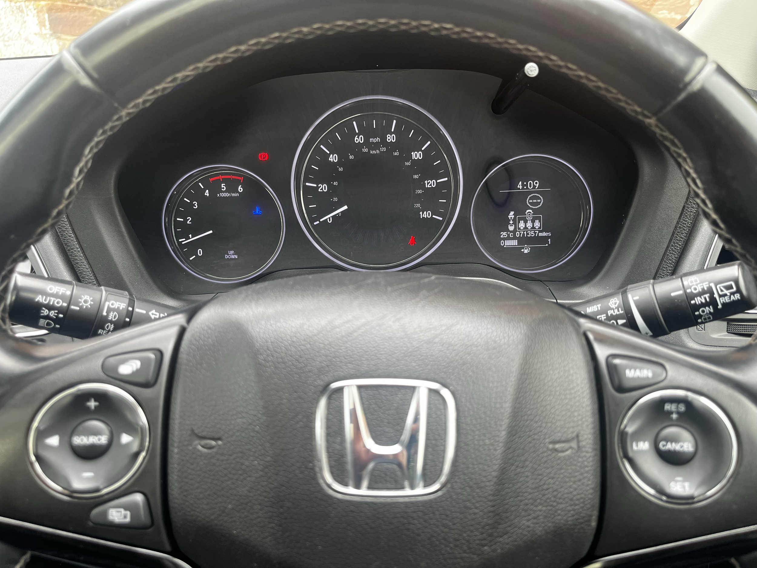 The image shows a car's dashboard from the driver's perspective, including speedometers, warning lights, and control stalks on either side of the steering wheel, with the Honda logo visible on the steering wheel.