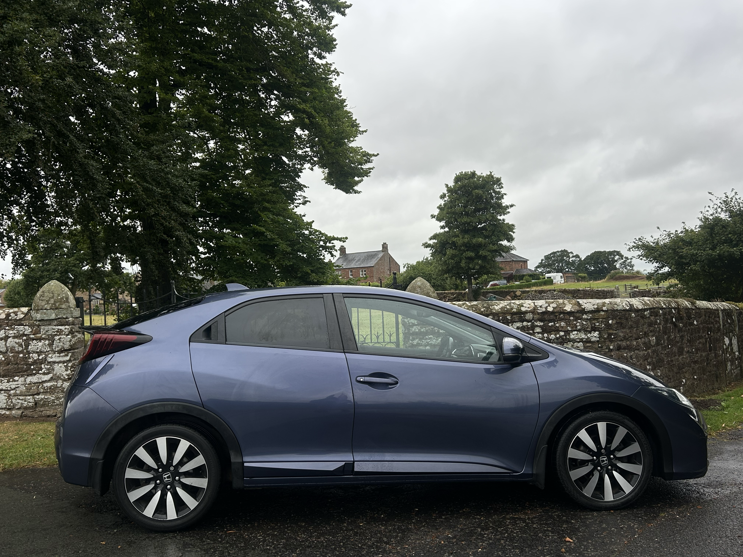 A blue Honda sedan parked beside a stone wall with trees and houses in the background on a cloudy day.