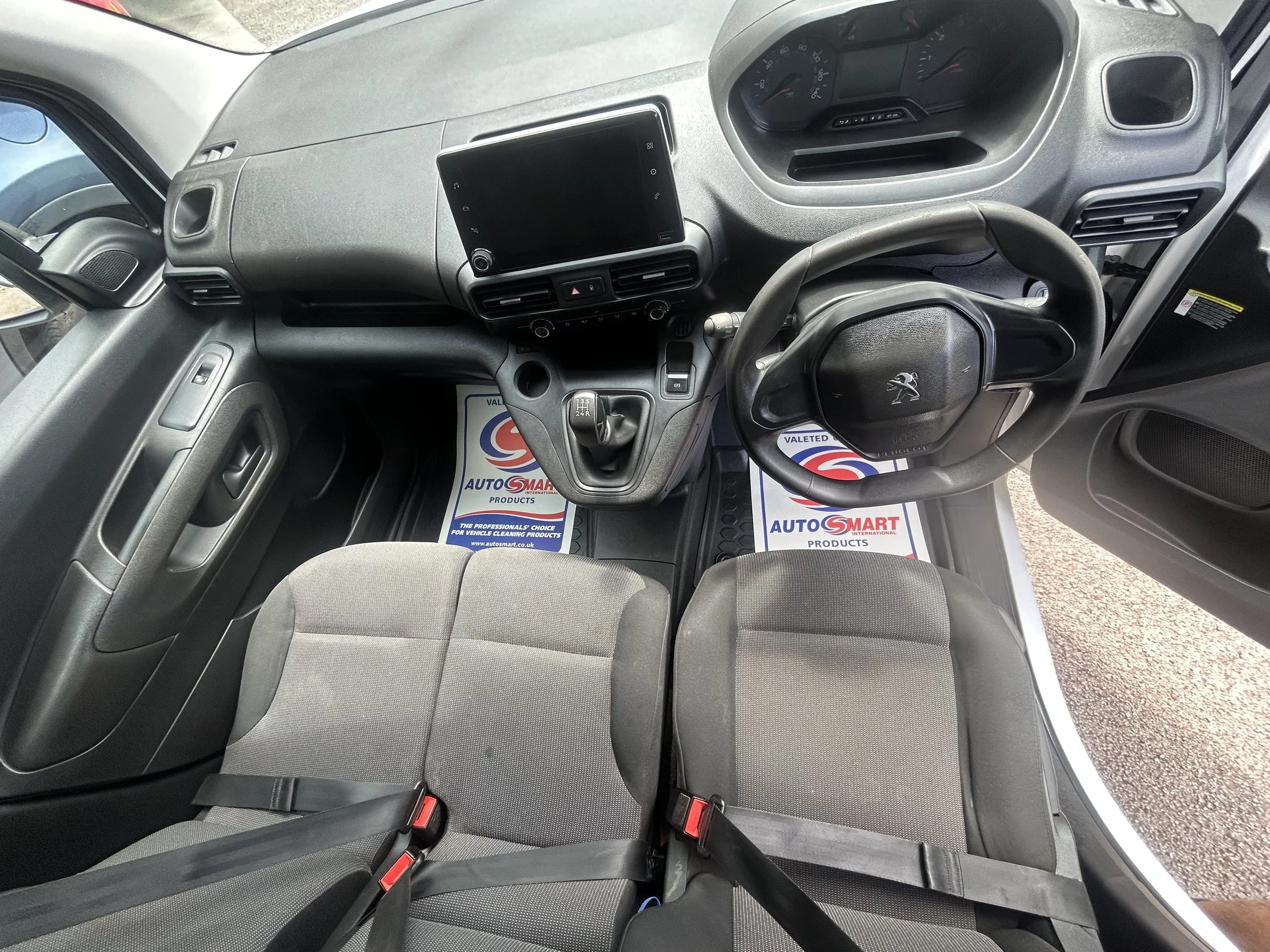Interior of a vehicle's front cabin showing two gray seats, a steering wheel with a logo, a gear shift, a dashboard, and a digital display screen.