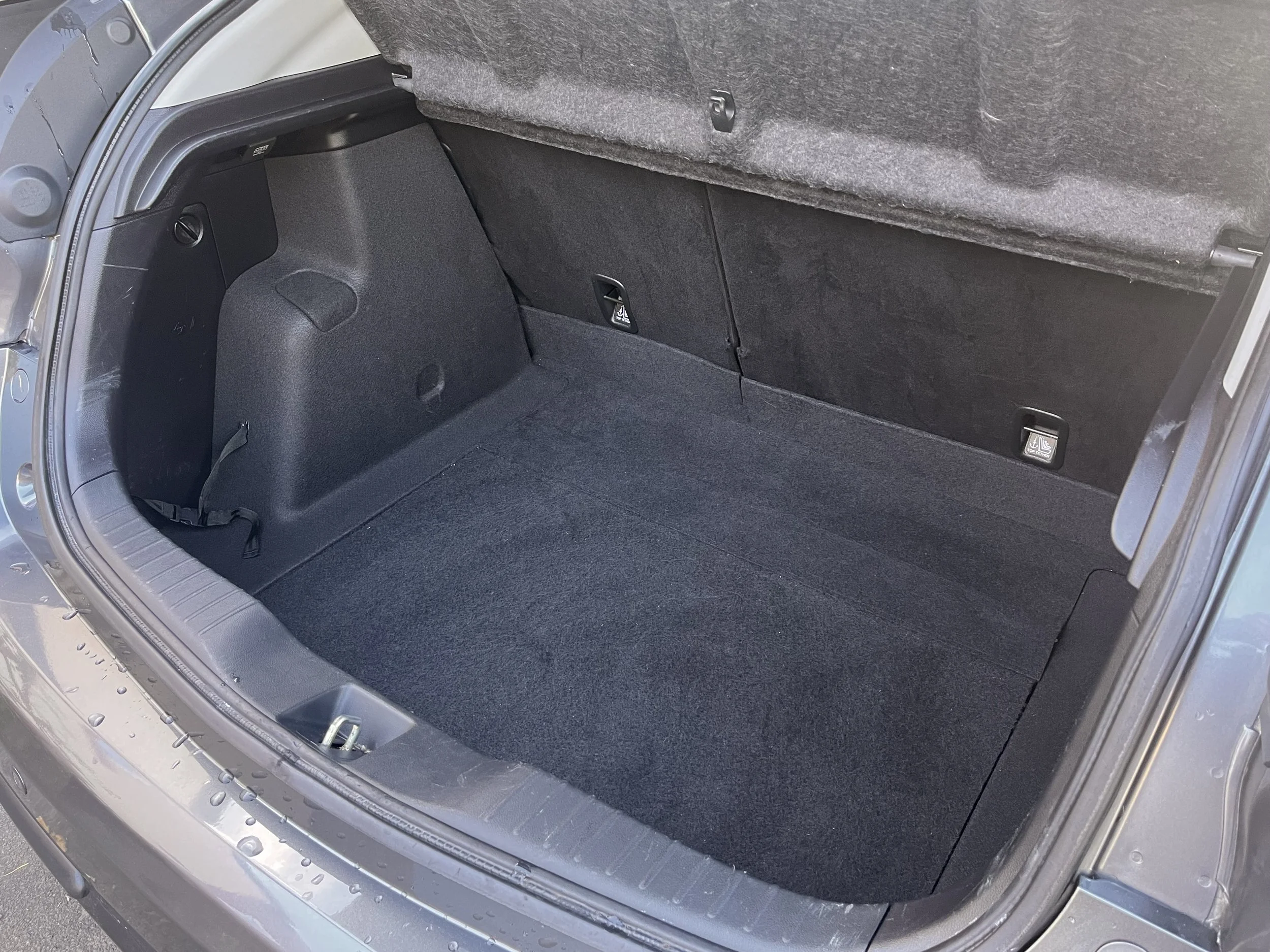 Empty car trunk with black interior carpeting and rear seats, showing latch points and a strap on the left side.