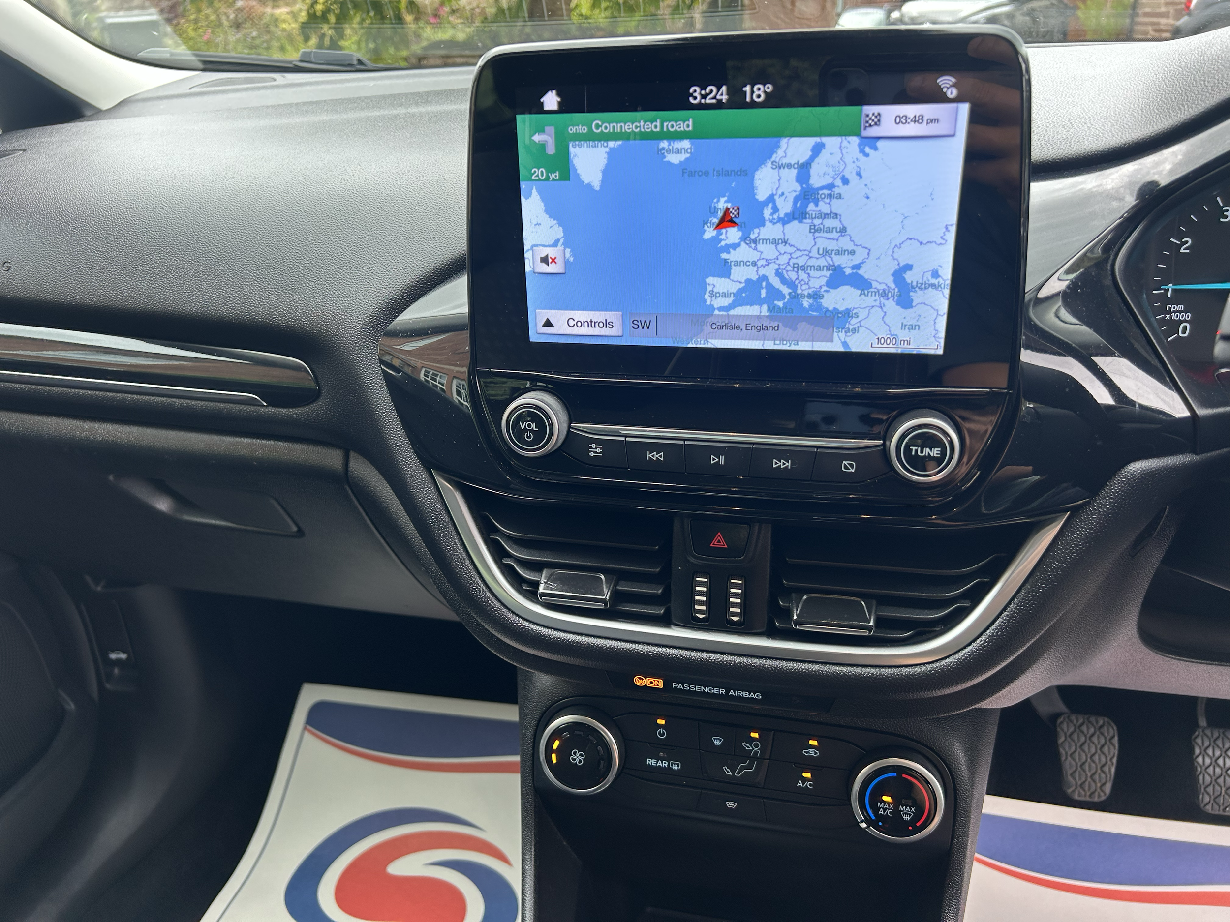 Car's dashboard with GPS navigation screen showing a map of Europe, with a route to Carlisle, England, and various controls and climate settings.