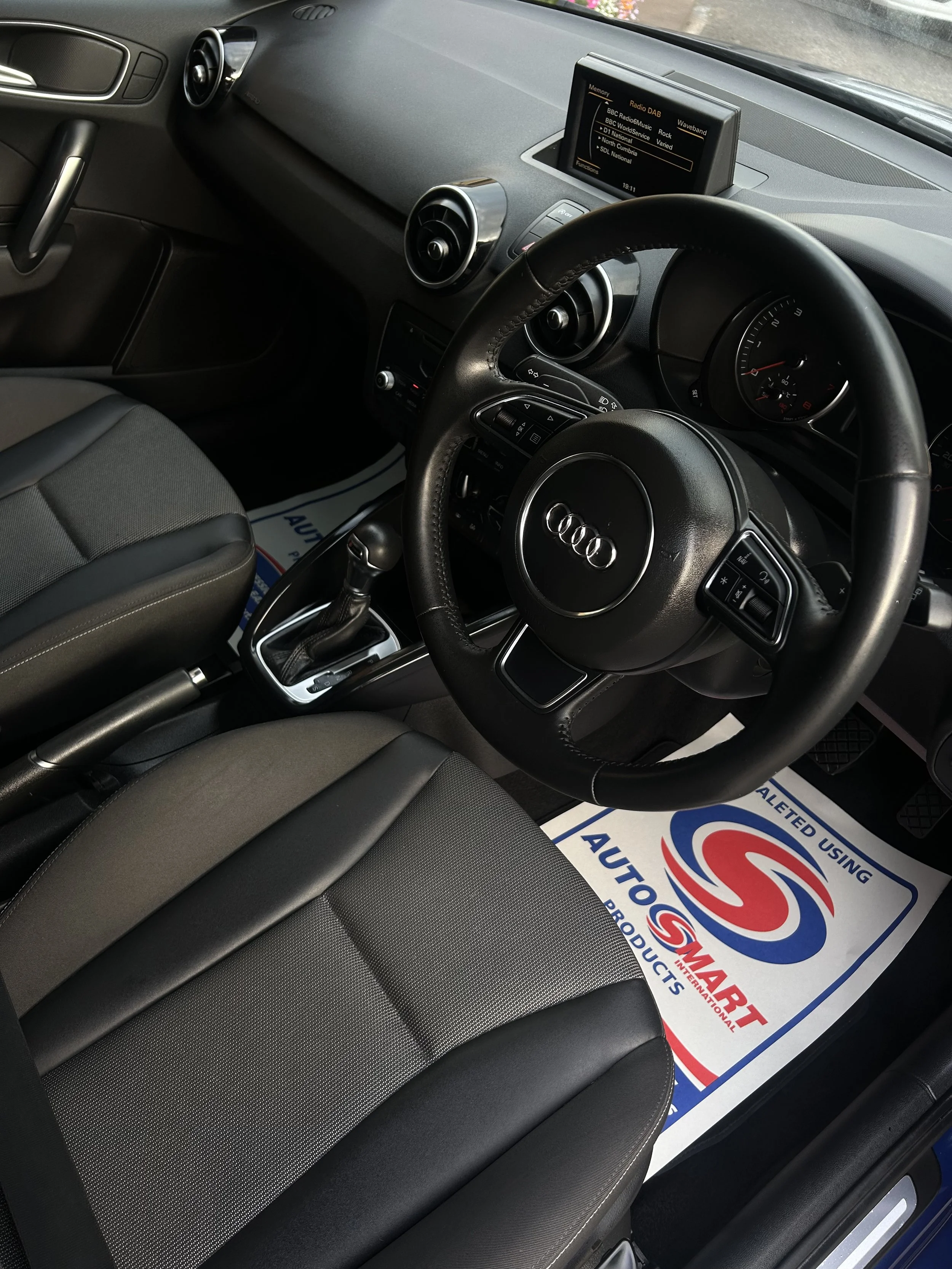 Interior of an Audi car, showing the steering wheel, dashboard, gear shift, driver seat, and a floor mat with the logo of Autocare.