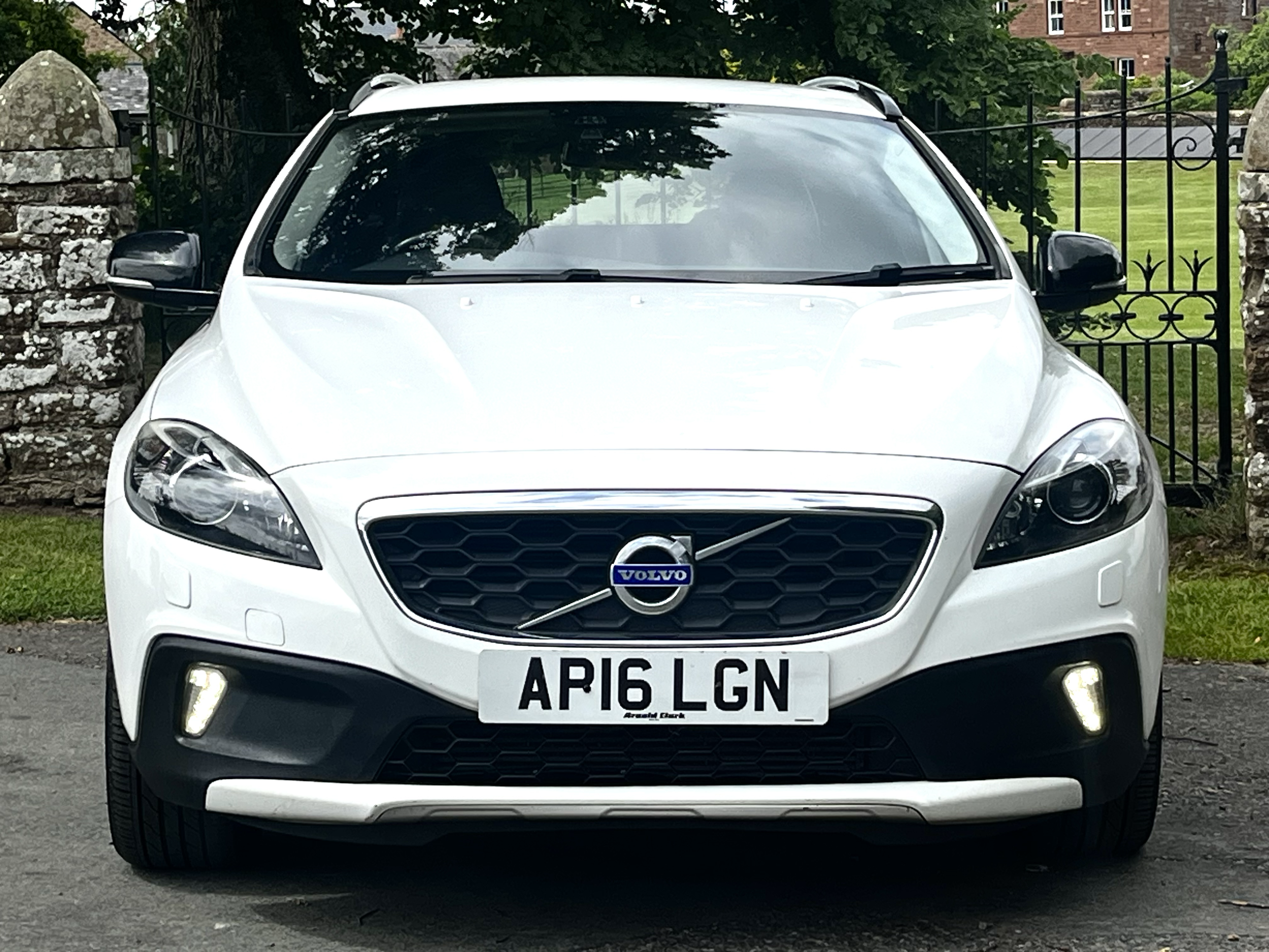 Front view of a white Volvo car with a black grille, displaying the registration plate 'AP16 LGN', parked in front of a stone wall and black wrought-iron gate, with trees and a grassy area in the background.