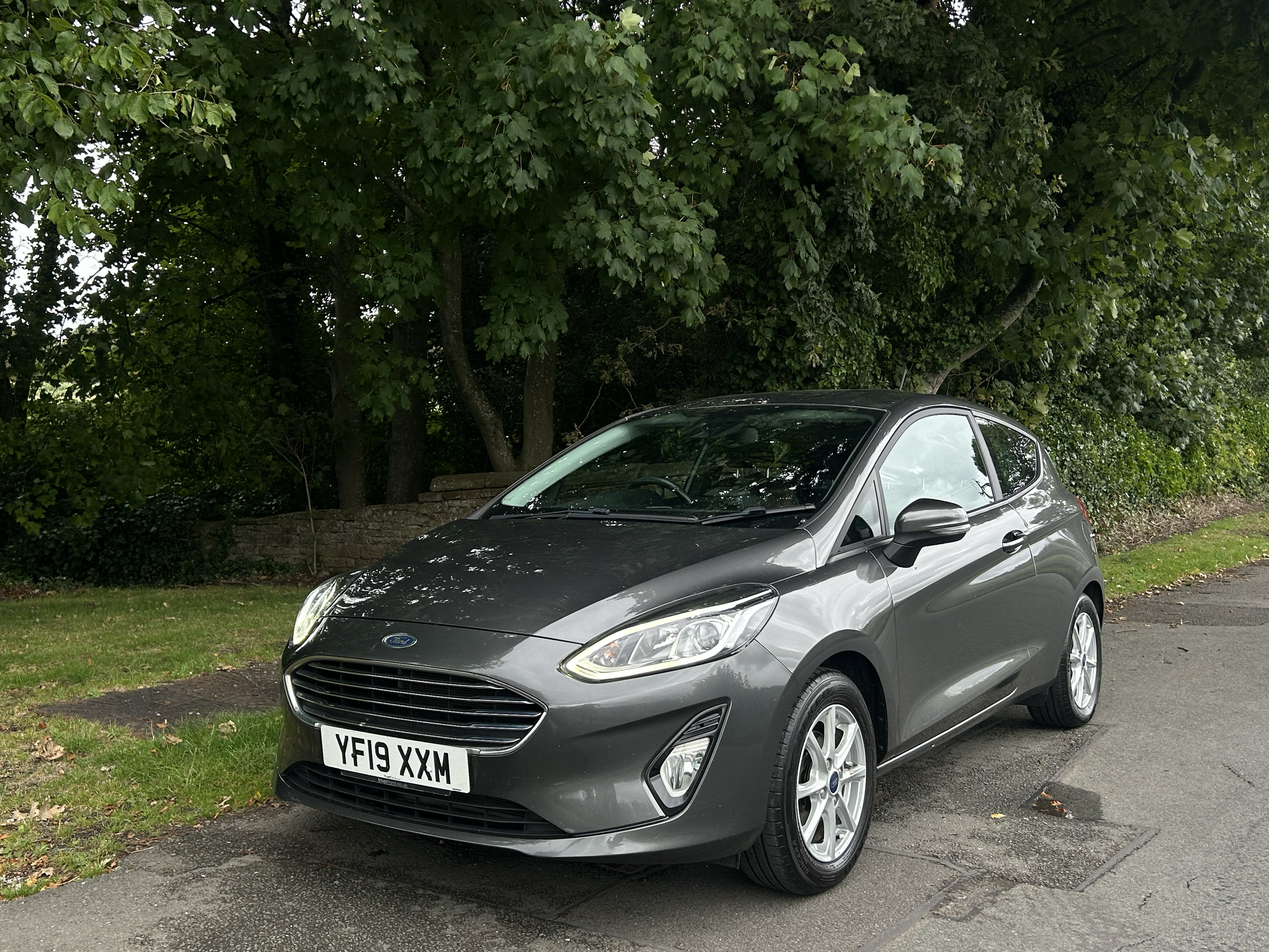 A gray Ford Fiesta parked on a paved driveway with green grass and trees in the background.