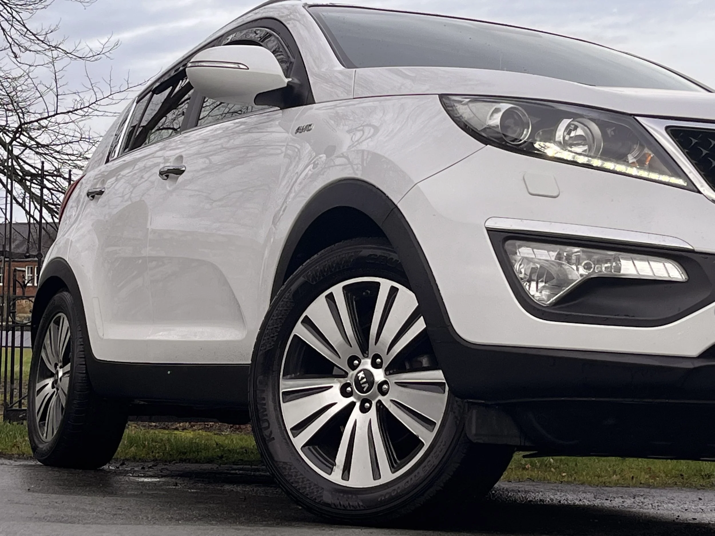 White Kia compact car parked on a street near a grassy area with trees in the background.