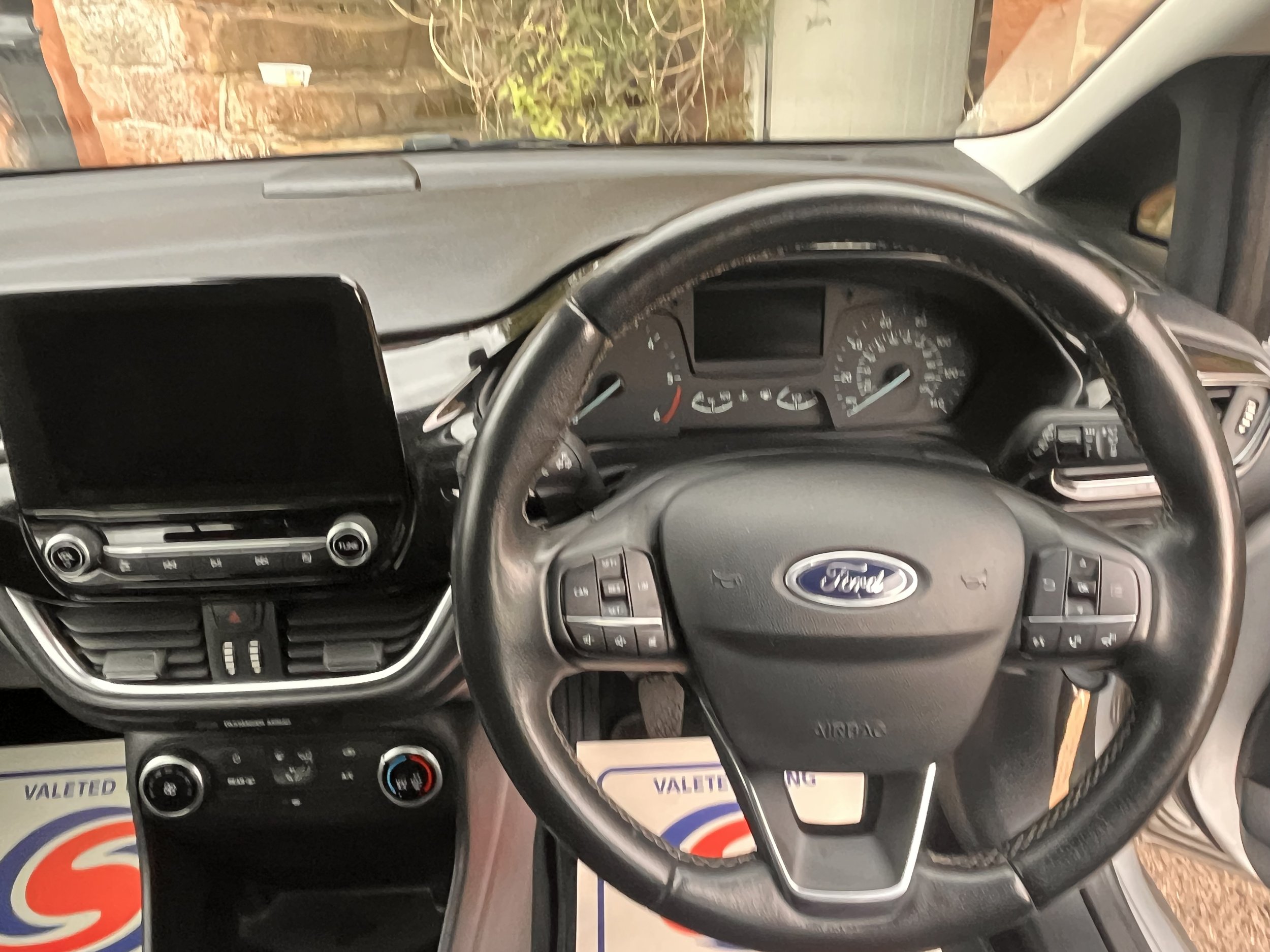 The interior of a Ford vehicle, showing the steering wheel with logo, dashboard with speedometer and other gauges, central console with touchscreen display, climate controls, and air vents.