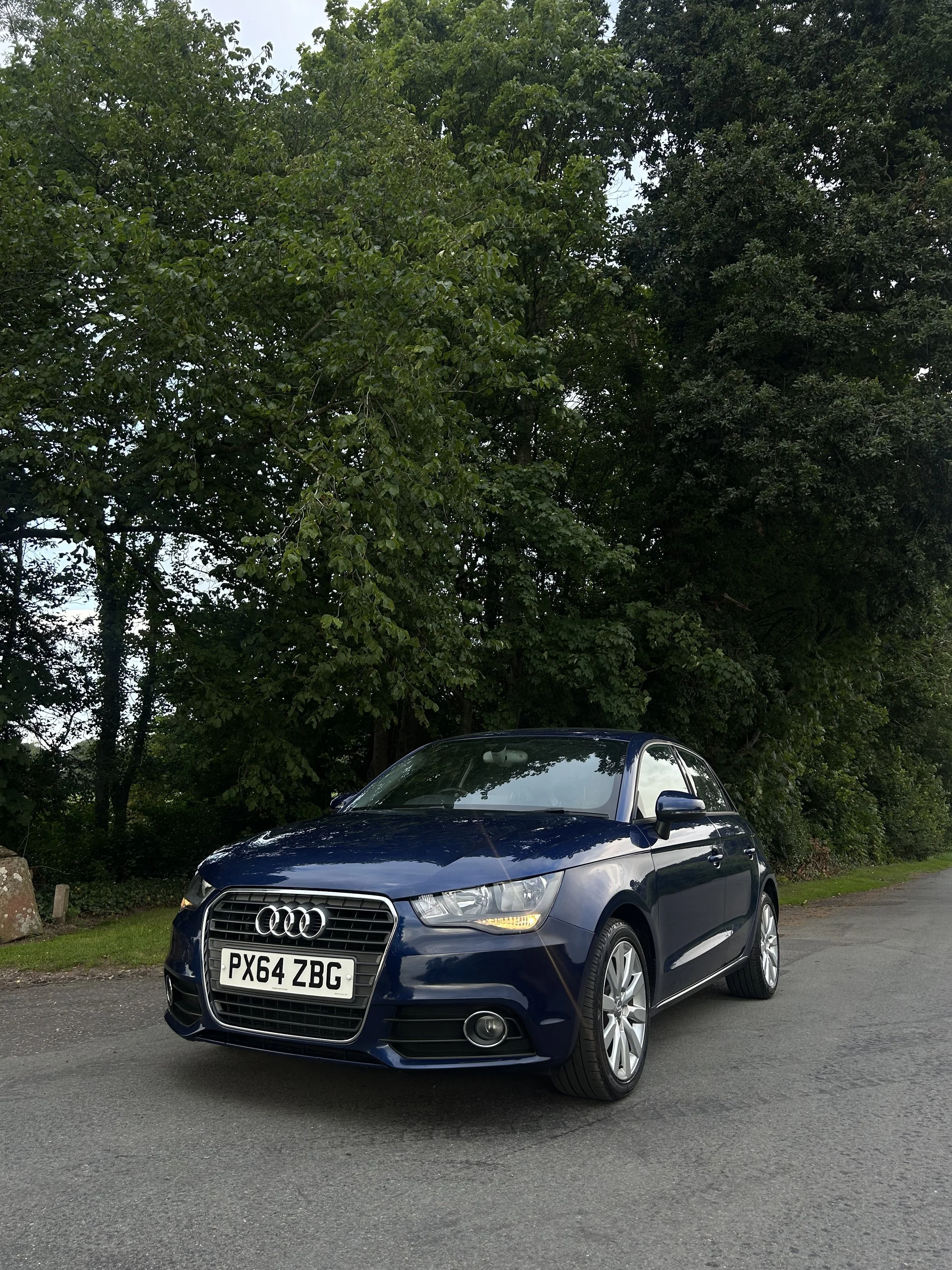 A blue Audi car parked on the side of a road with trees in the background.