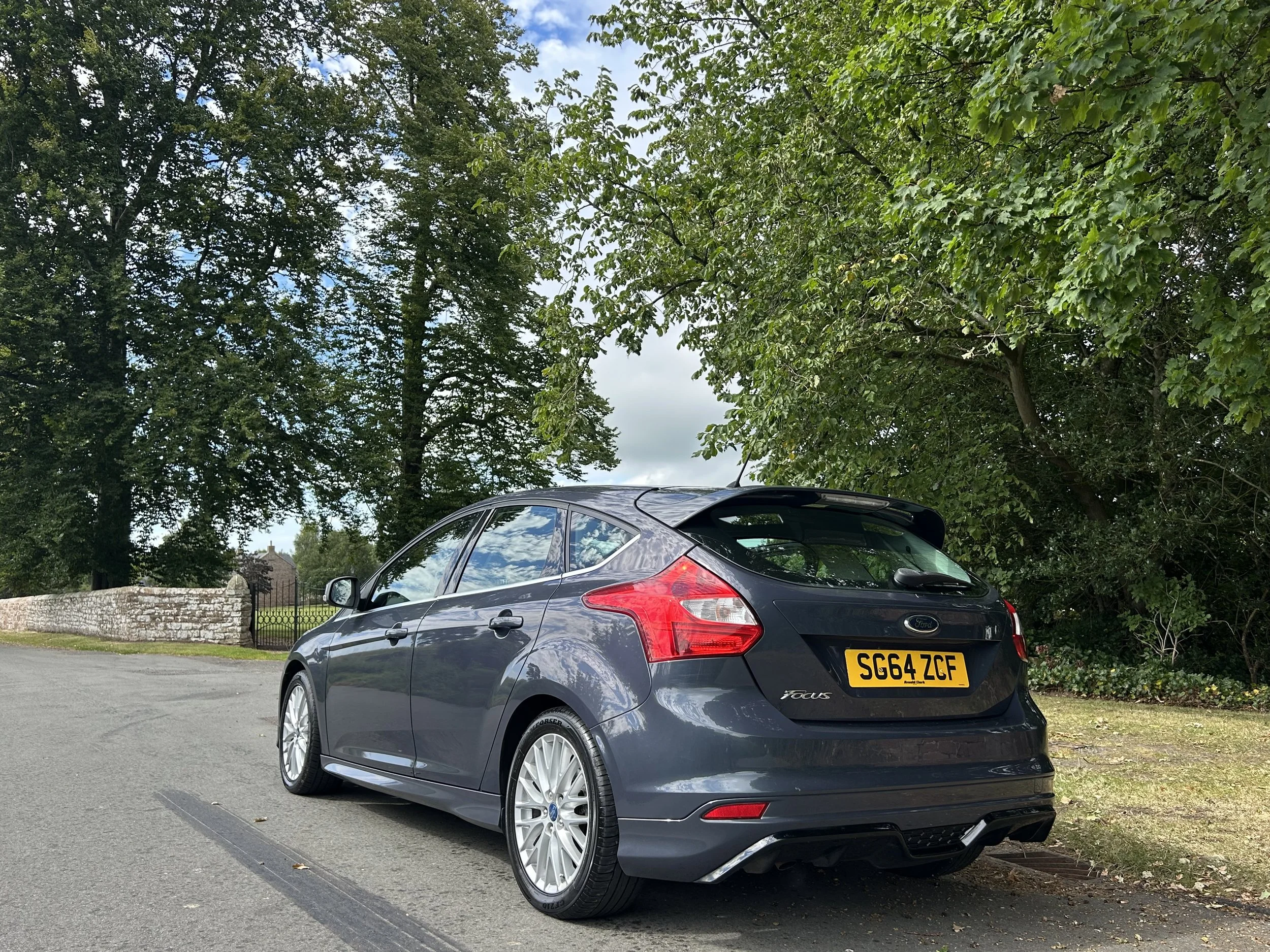 A dark gray Ford Focus hatchback parked on the side of a road with green trees and a stone wall in the background.
