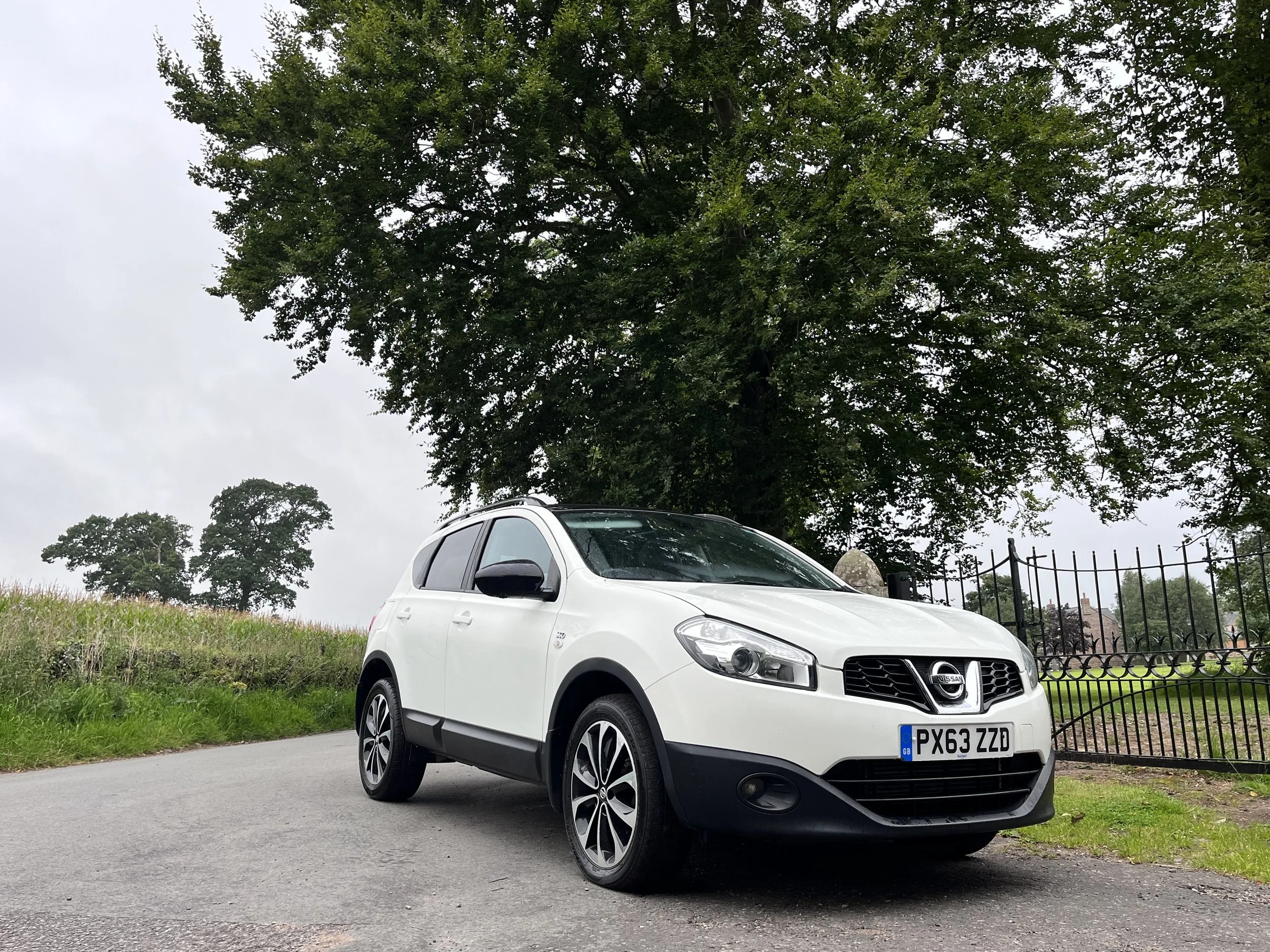 White Nissan SUV parked on a paved road with grass and trees in the background under cloudy skies.