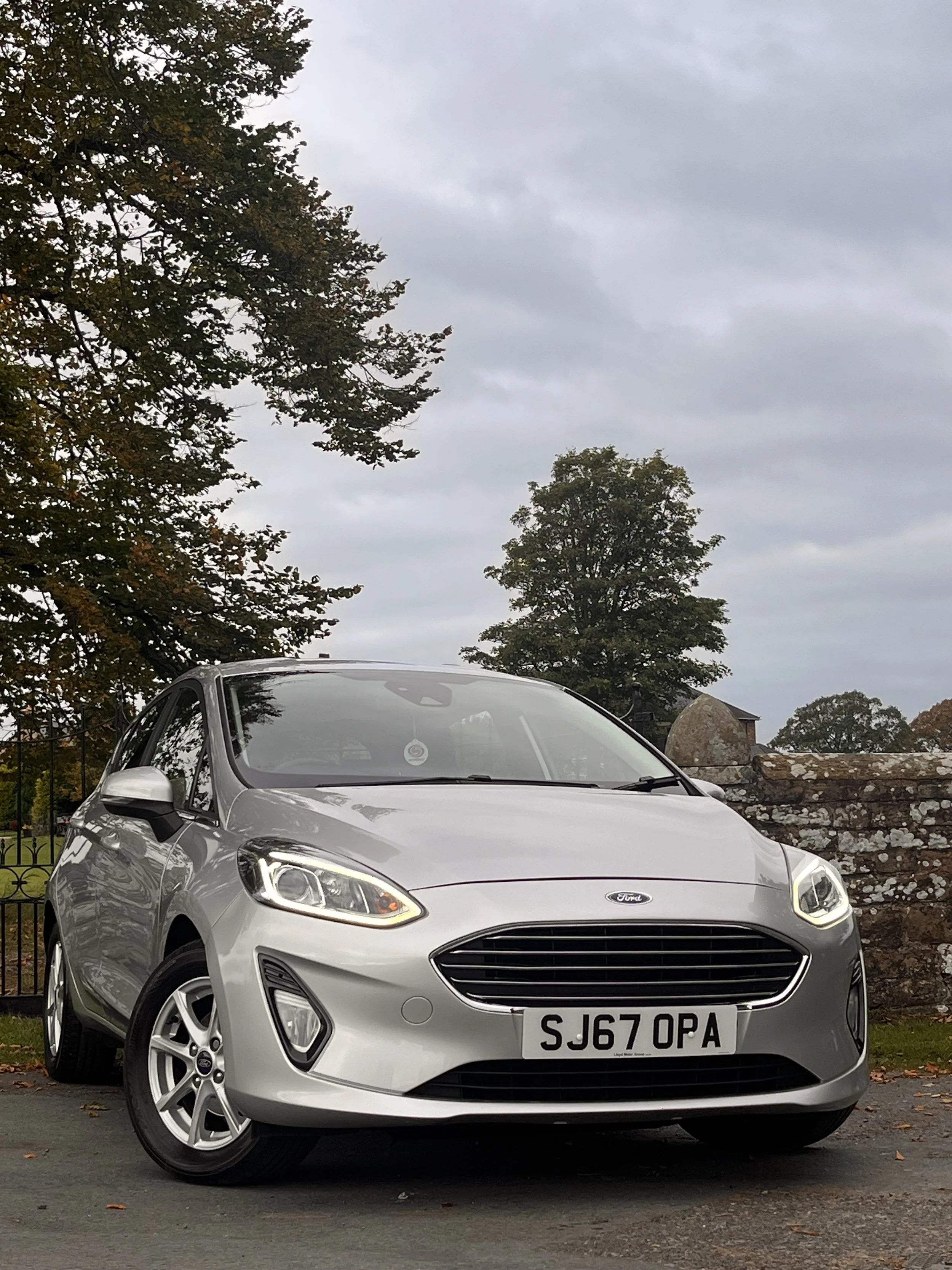 Silver Ford Fiesta parked outdoors on a cloudy day with trees and a stone wall in the background.