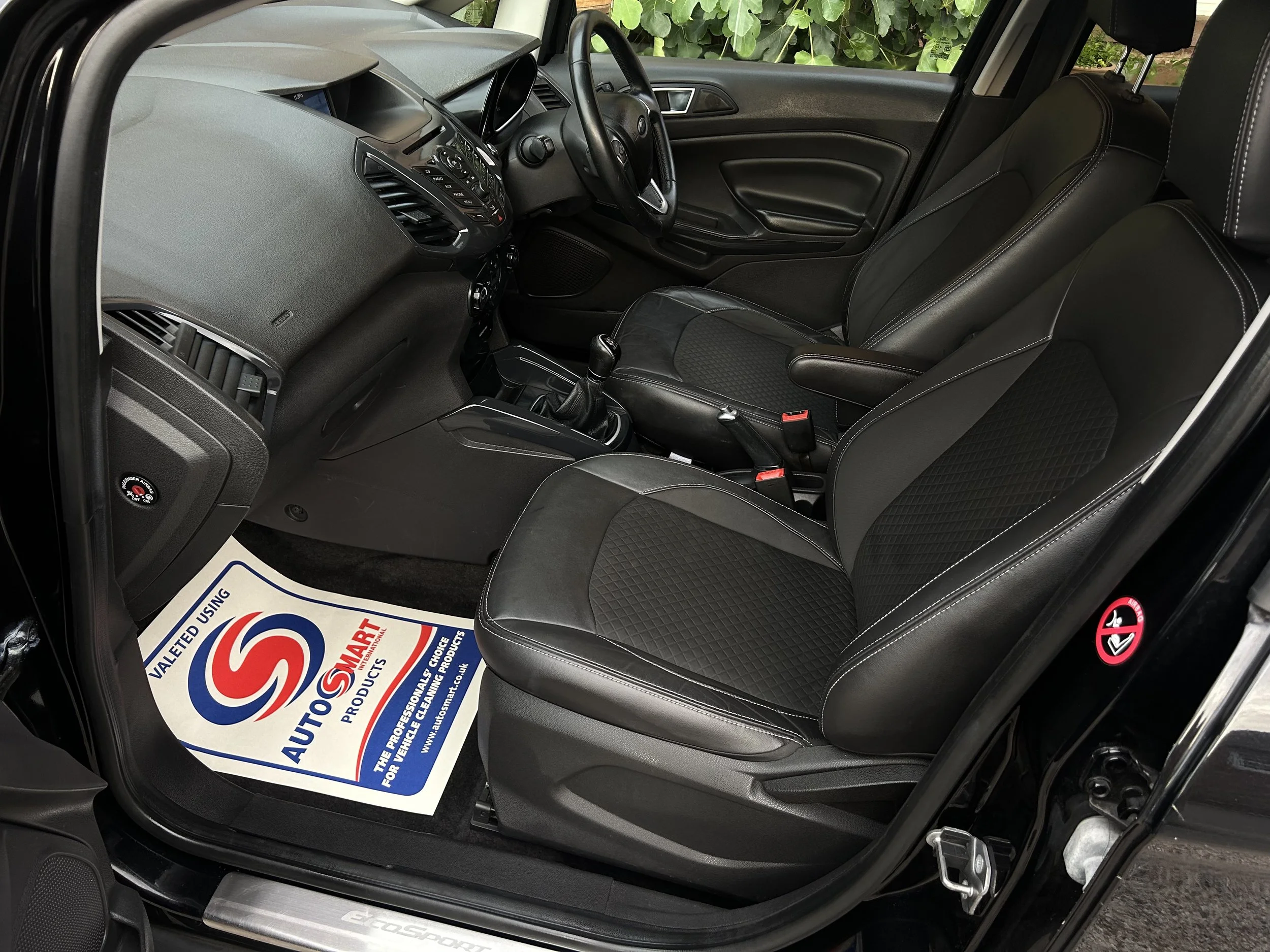The interior of a black car showing the front seats, dashboard, steering wheel, and a valeted mat on the driver's side.