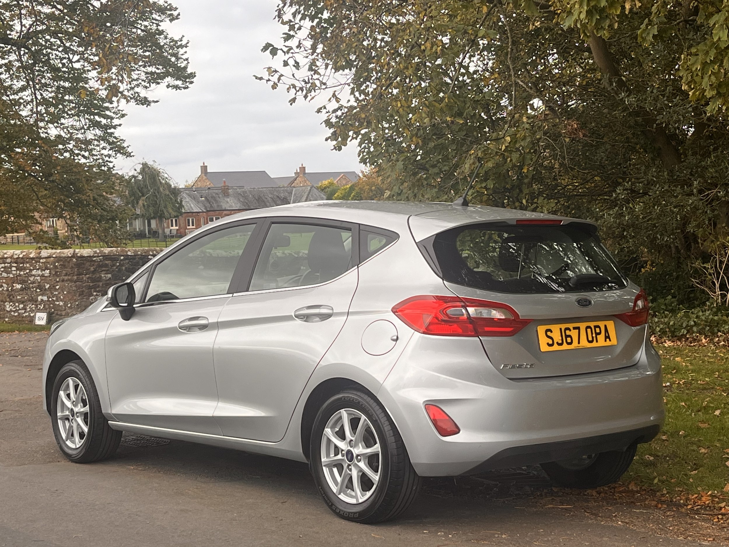 Silver hatchback car parked on the side of the road with trees and a stone wall in the background.