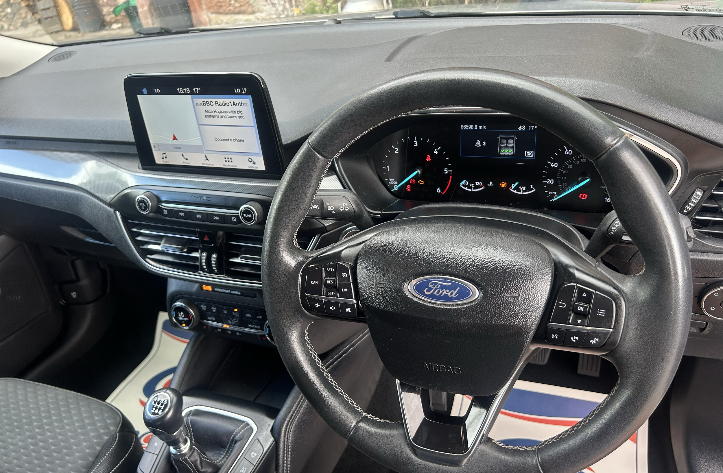 View of car dashboard showing steering wheel, instrument panel with speedometer, tachometer, and warning lights, and in-car multimedia display with BBC Radio 1 Anthem playing. Driver's hand on steering wheel.