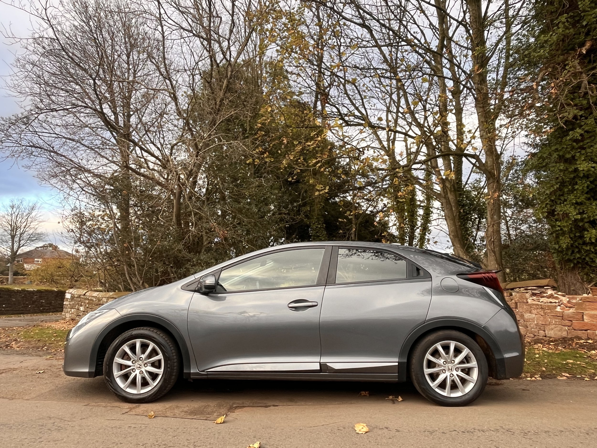 A gray Honda Civic parked on a street during autumn with fallen leaves, leafless trees, and a brick wall in the background.
