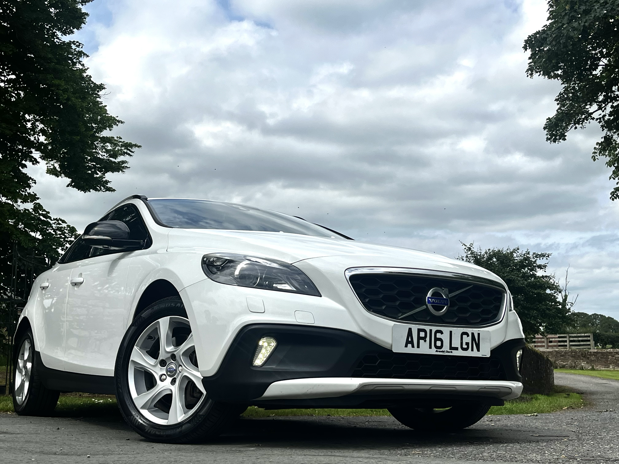 White Volvo SUV car parked outdoors under a cloudy sky with trees in the background and a partial view of the front and side of the vehicle.
