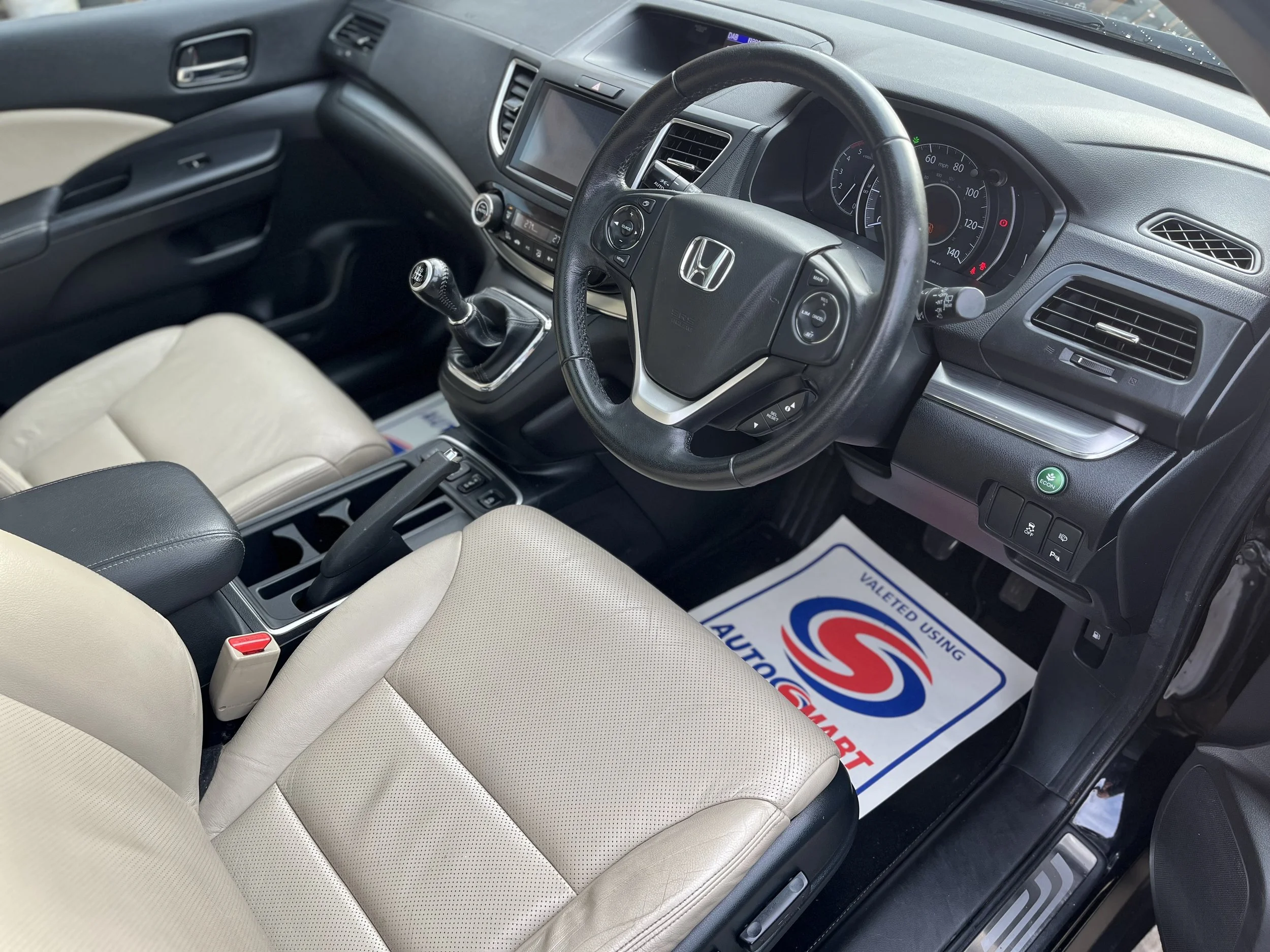 Interior view of a Honda car showing the steering wheel, gear shift, dashboard, front seats with light-colored upholstery, and a floor mat with the 'Autosmart' logo.