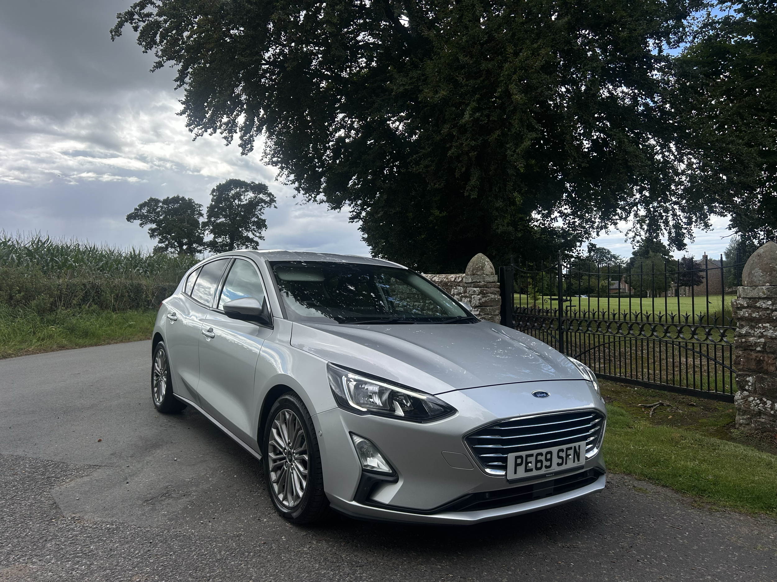 A silver Ford car with the license plate PE69 SFN parked on a paved road in front of a black metal fence and stone pillars, with large trees and a cloudy sky in the background.