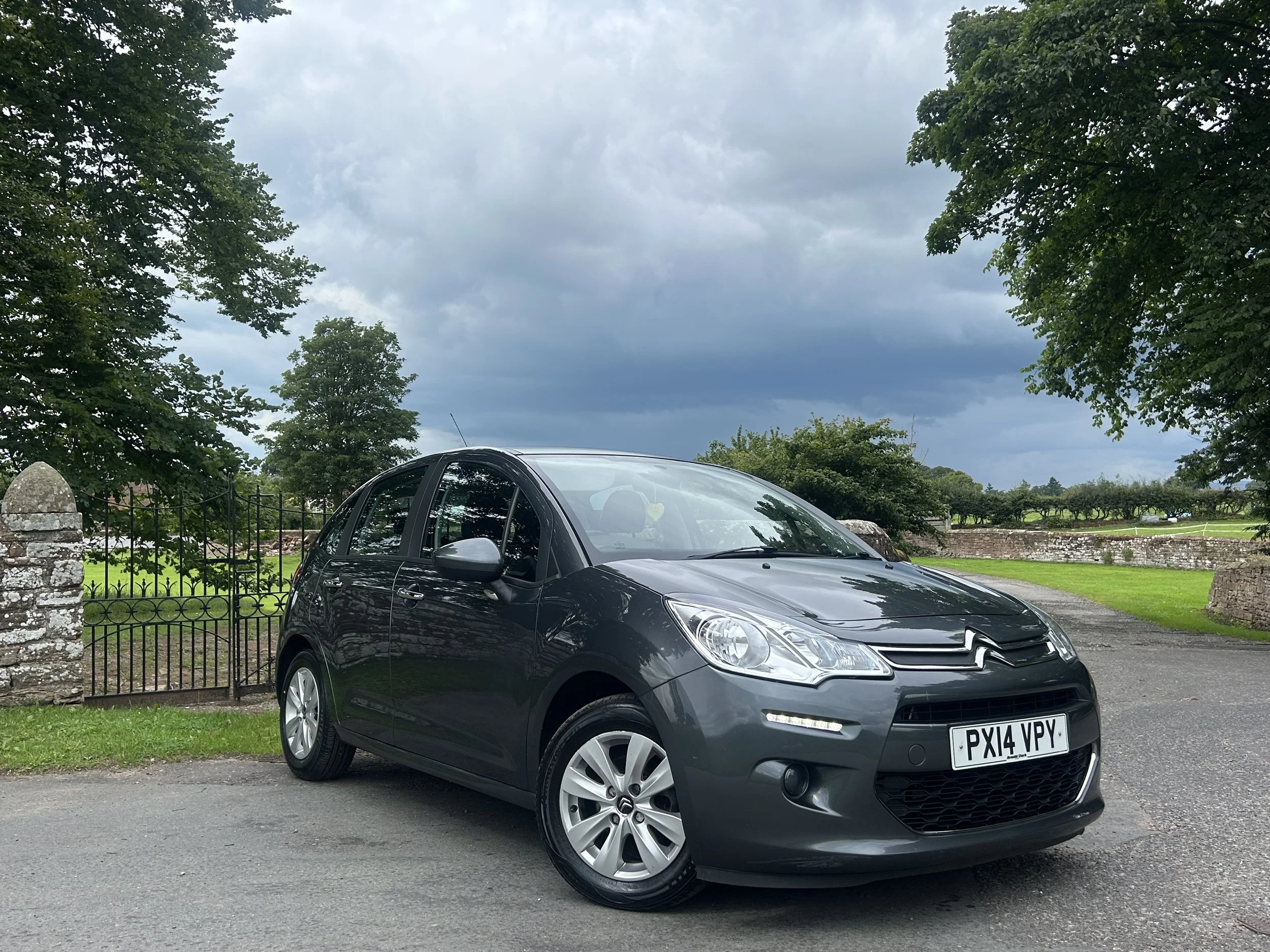 A black Citroën hatchback parked on a paved surface with a stone fence and green trees in the background under a cloudy sky.