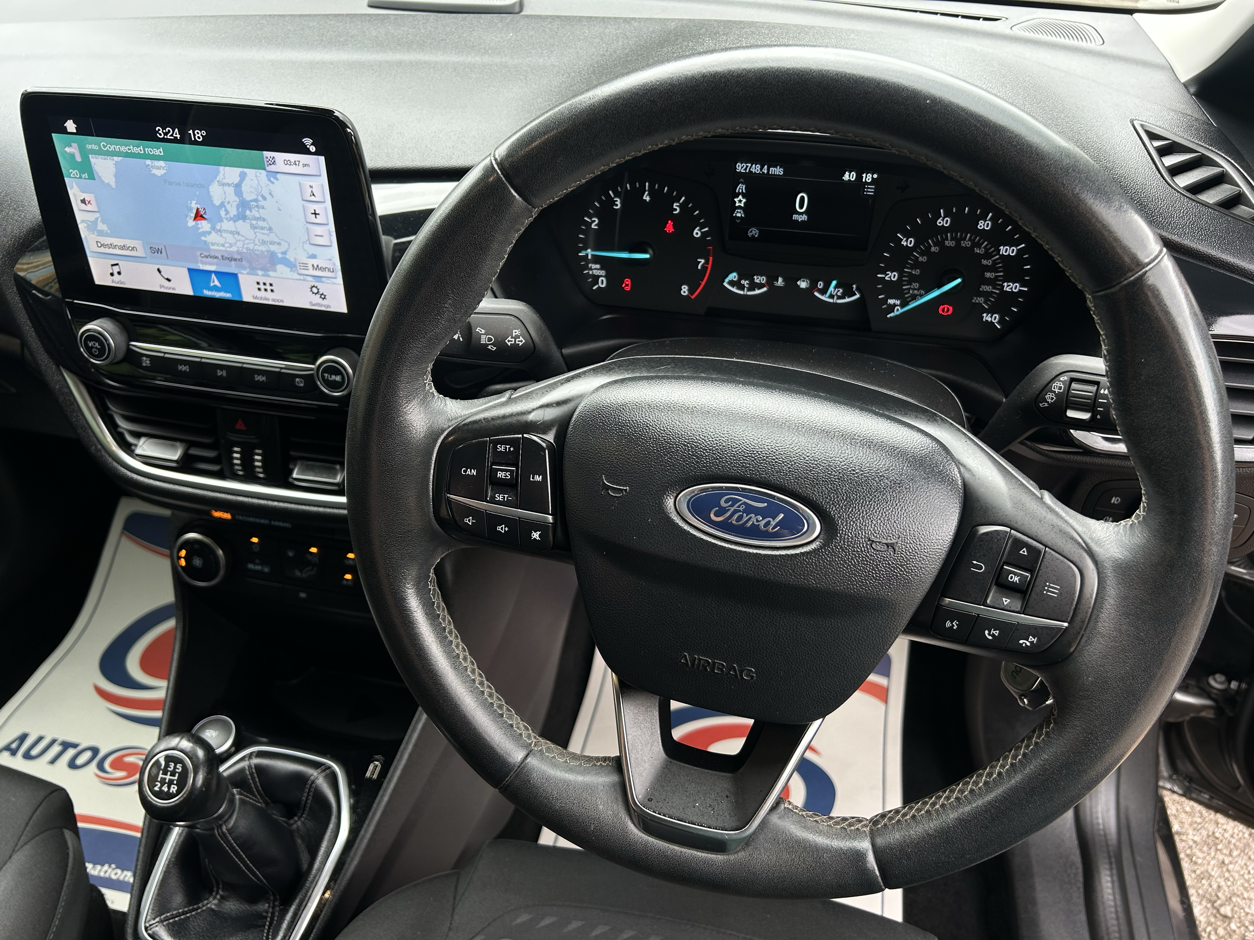 The image shows the interior of a Ford vehicle, including the steering wheel, dashboard, and infotainment screen displaying a map. The gear shift lever and various controls are also visible.