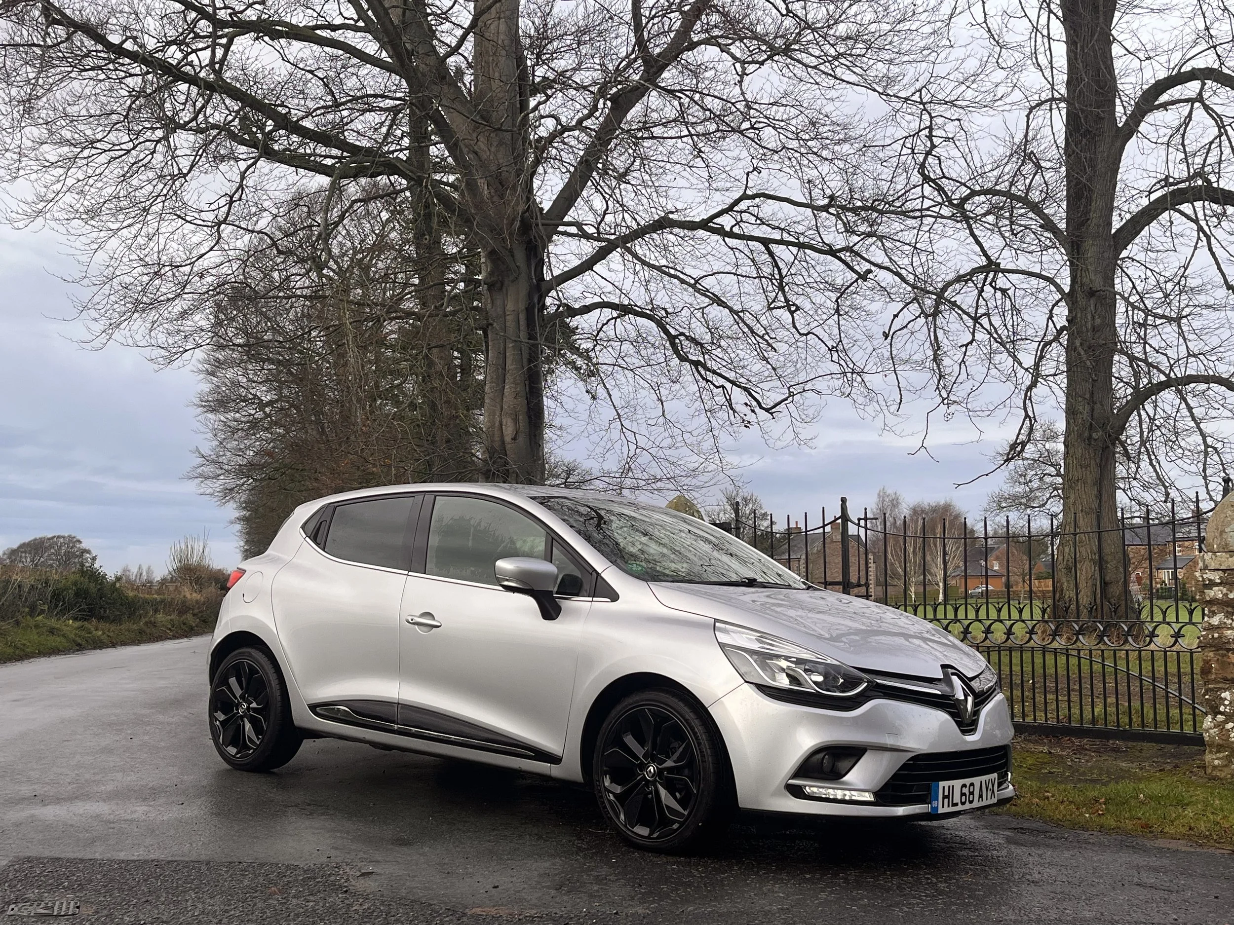 Silver Renault hatchback car parked on a wet road in front of a large tree and a black iron fence with houses and trees in the background under a cloudy sky.