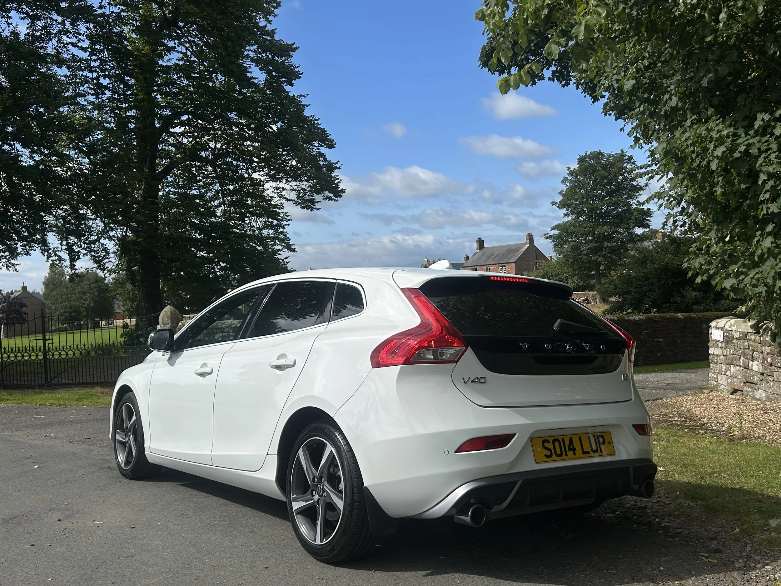 A white Volvo V40 hatchback parked on a street with trees, houses, and a brick wall in the background under a partly cloudy sky.