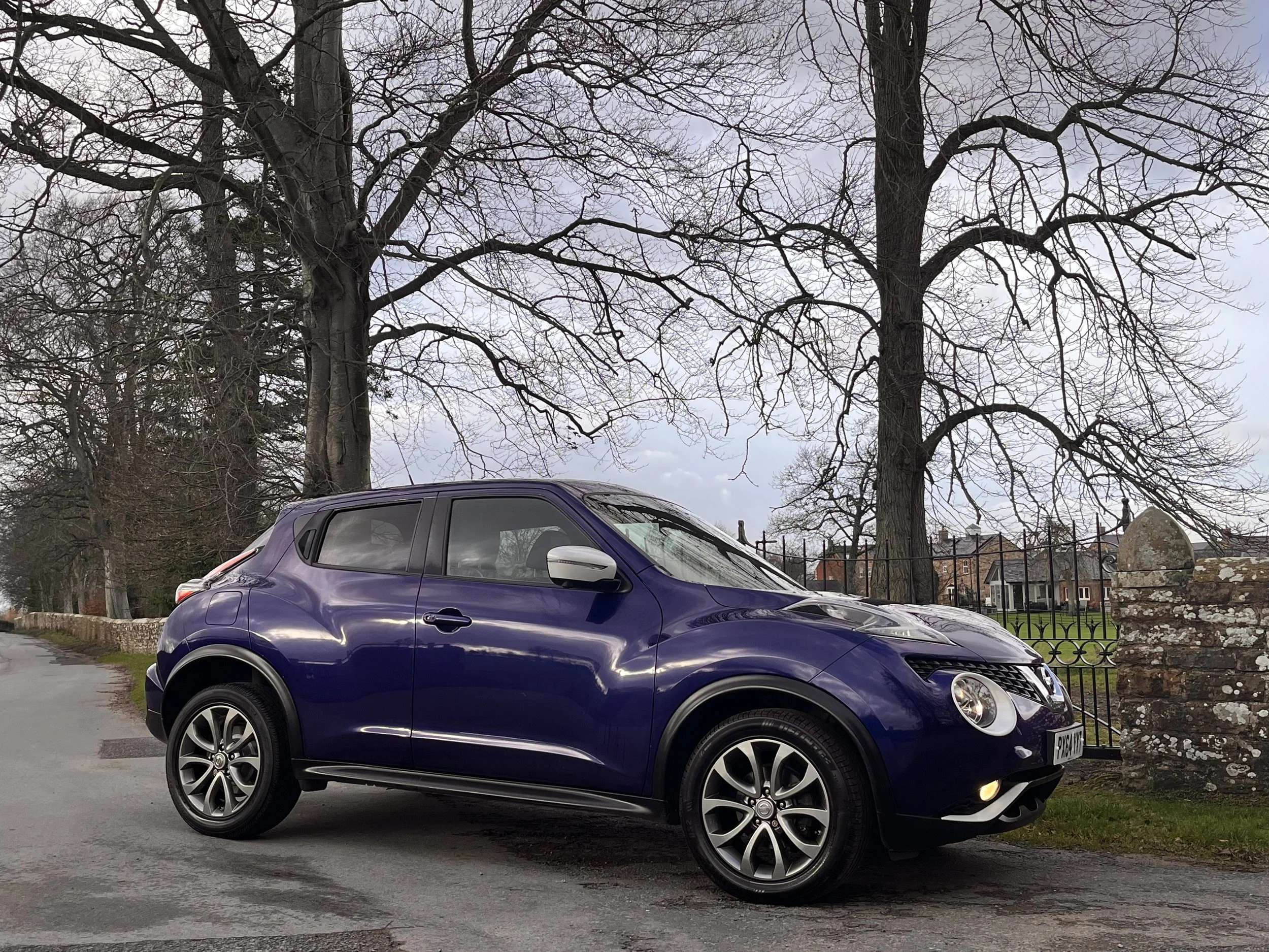 A purple Nissan Juke parked on a street with leafless trees and a stone fence in the background.