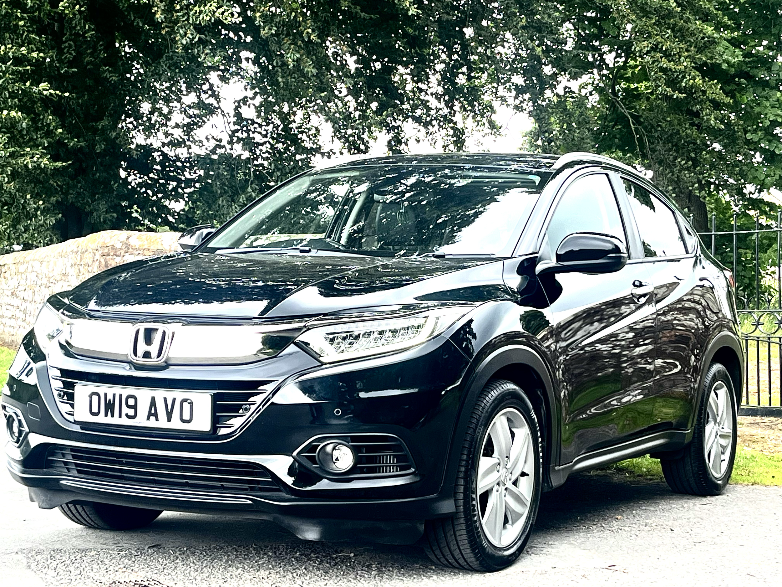 Black Honda HR-V parked on a gravel driveway, surrounded by green trees and a metal fence in the background.
