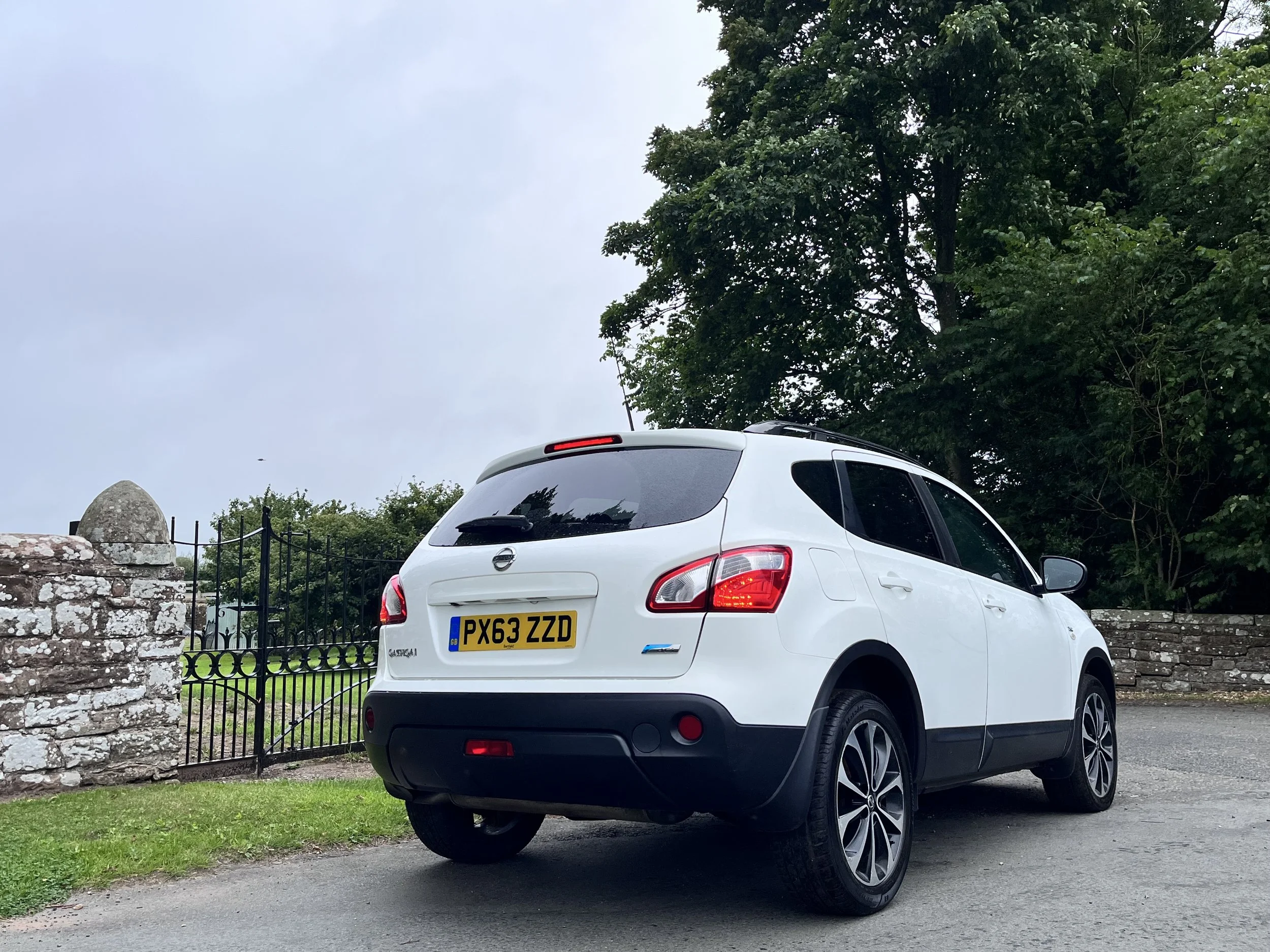 A white Nissan Qashqai parked on a gravel driveway near a stone wall and metal fence, with trees in the background and an overcast sky.
