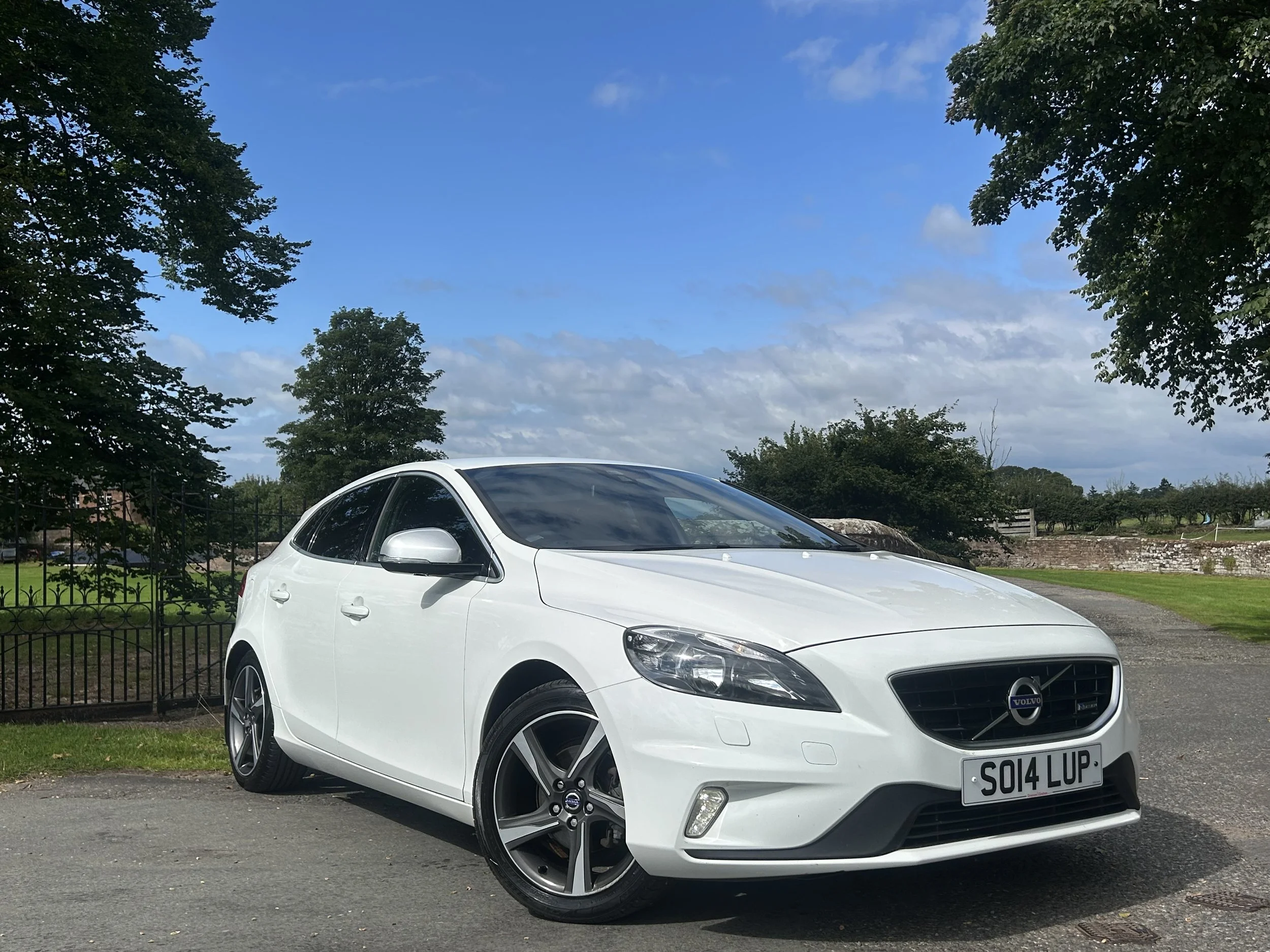 A white Volvo hatchback car parked on a paved surface near a garden with green grass, trees, and a fence, under a partly cloudy blue sky.