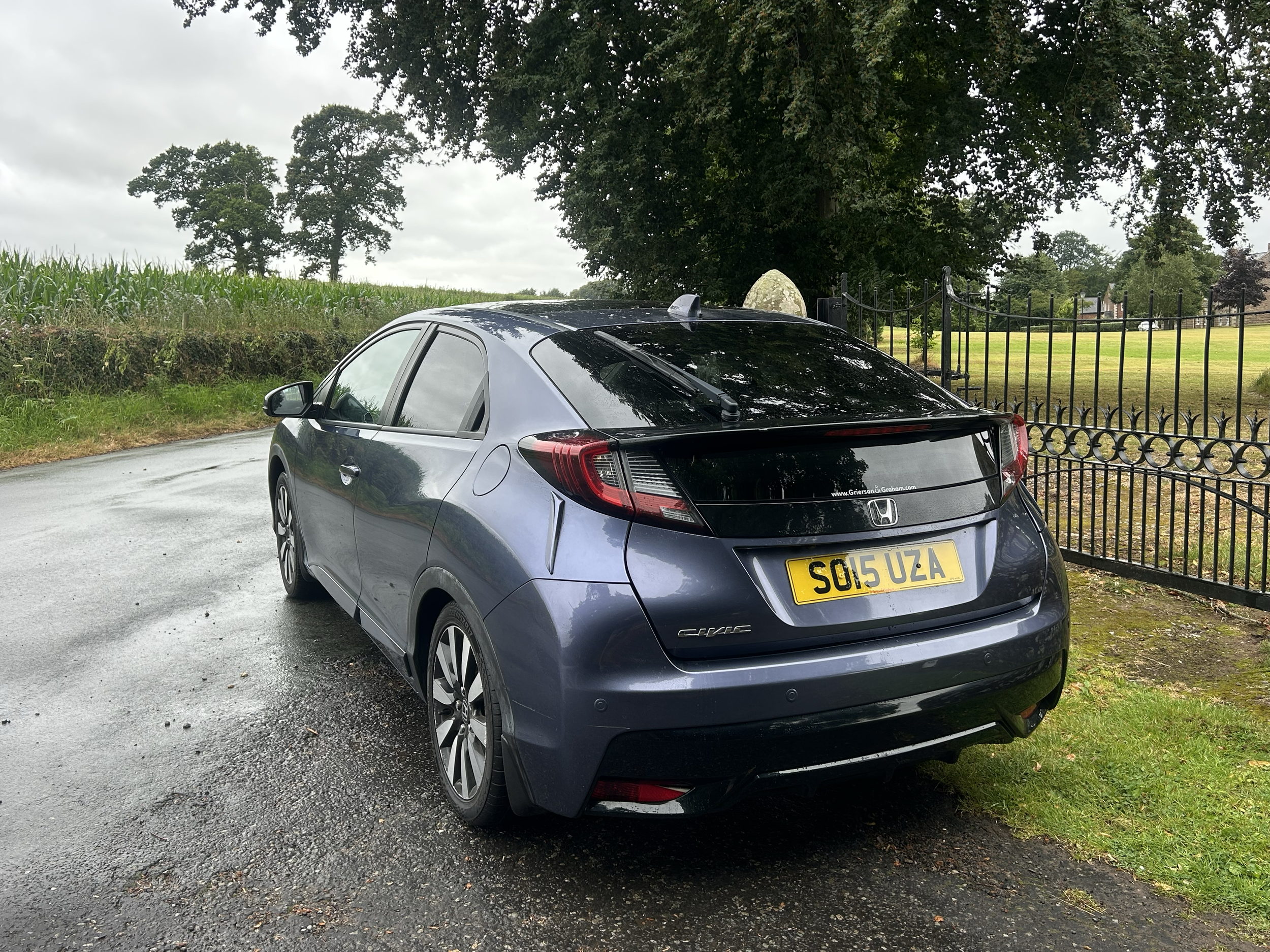 A gray Honda Civic parked on a wet road near a black fence, with trees and a grassy field in the background.