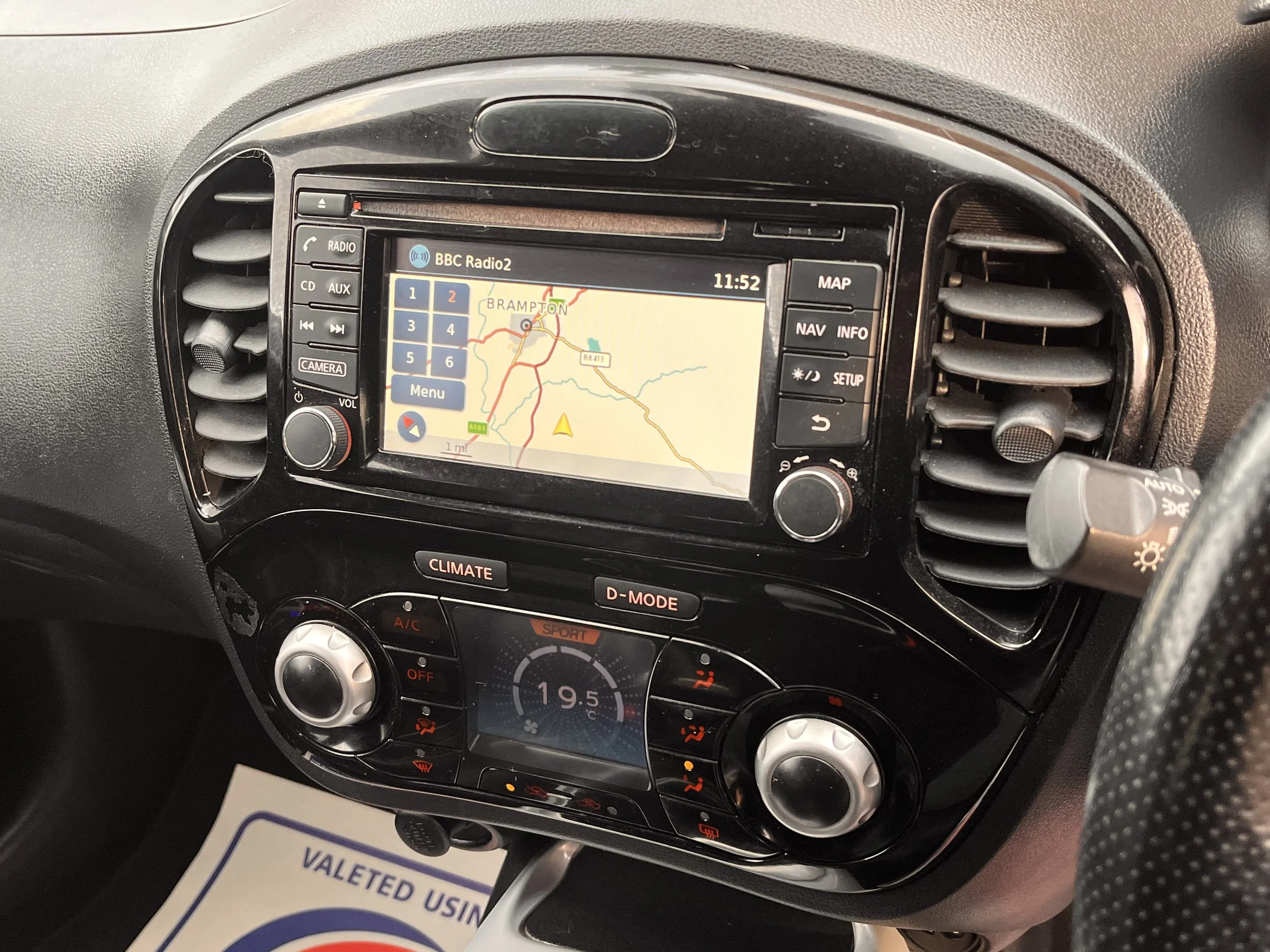 Car dashboard with navigation screen showing a map, climate controls set to 19.5°C, and radio tuned to BBC Radio 2, with air vents on either side.
