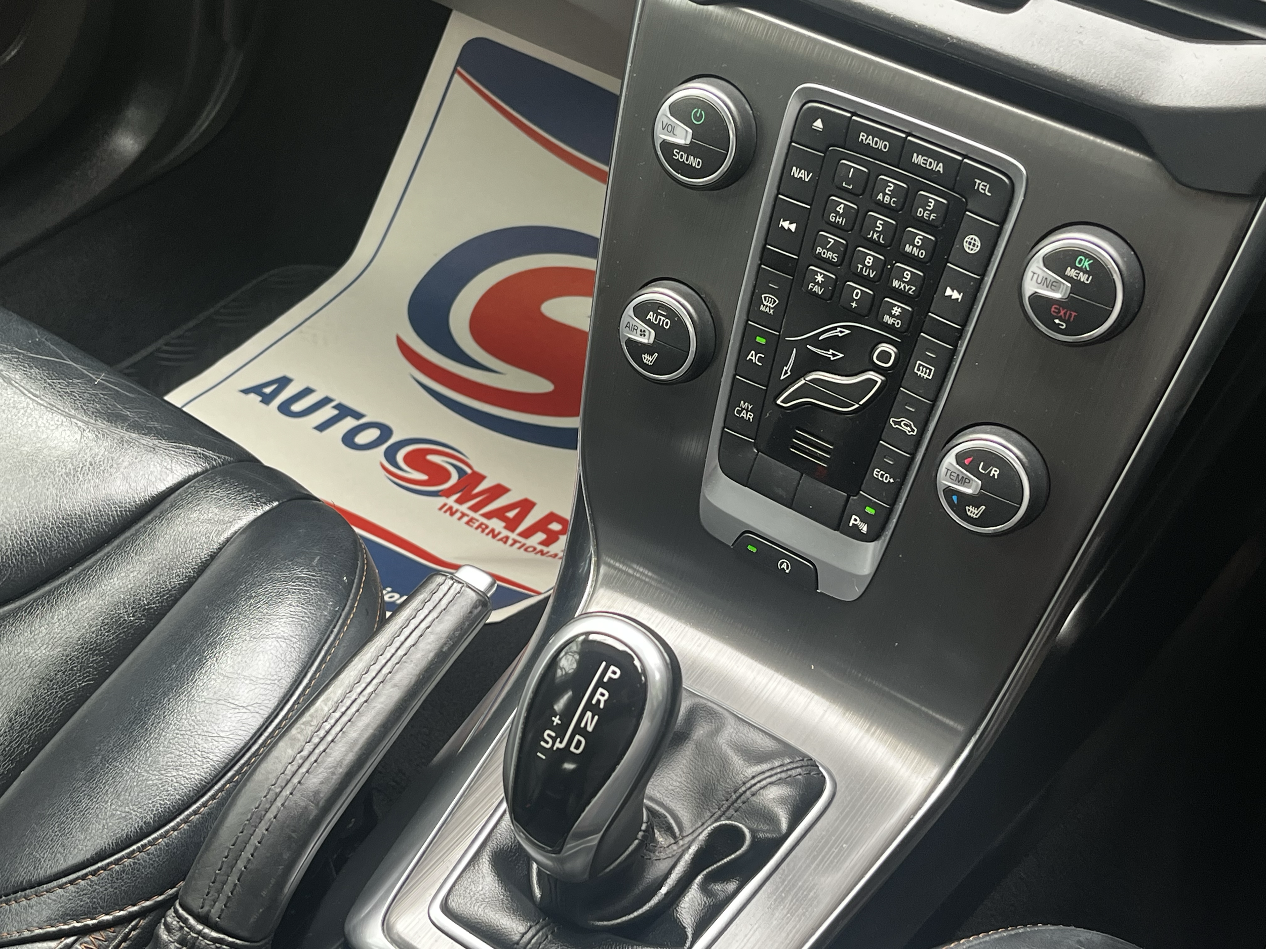 Close-up of a car's center console showing controls for audio, navigation, and climate, with a gear shift and a black leather armrest in the foreground, and a floor mat with a logo in the background.